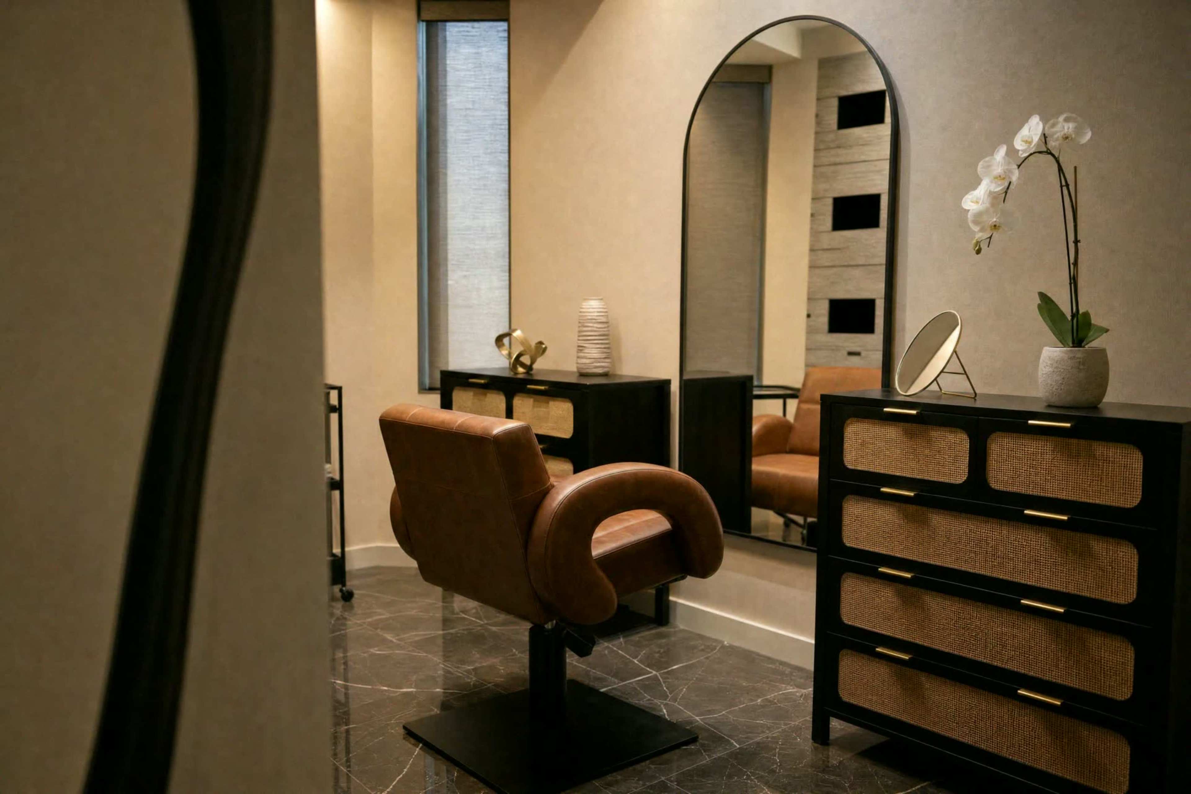 The image shows a modern salon interior featuring a leather salon chair, a round mirror, and a stylish dresser with decorative items.