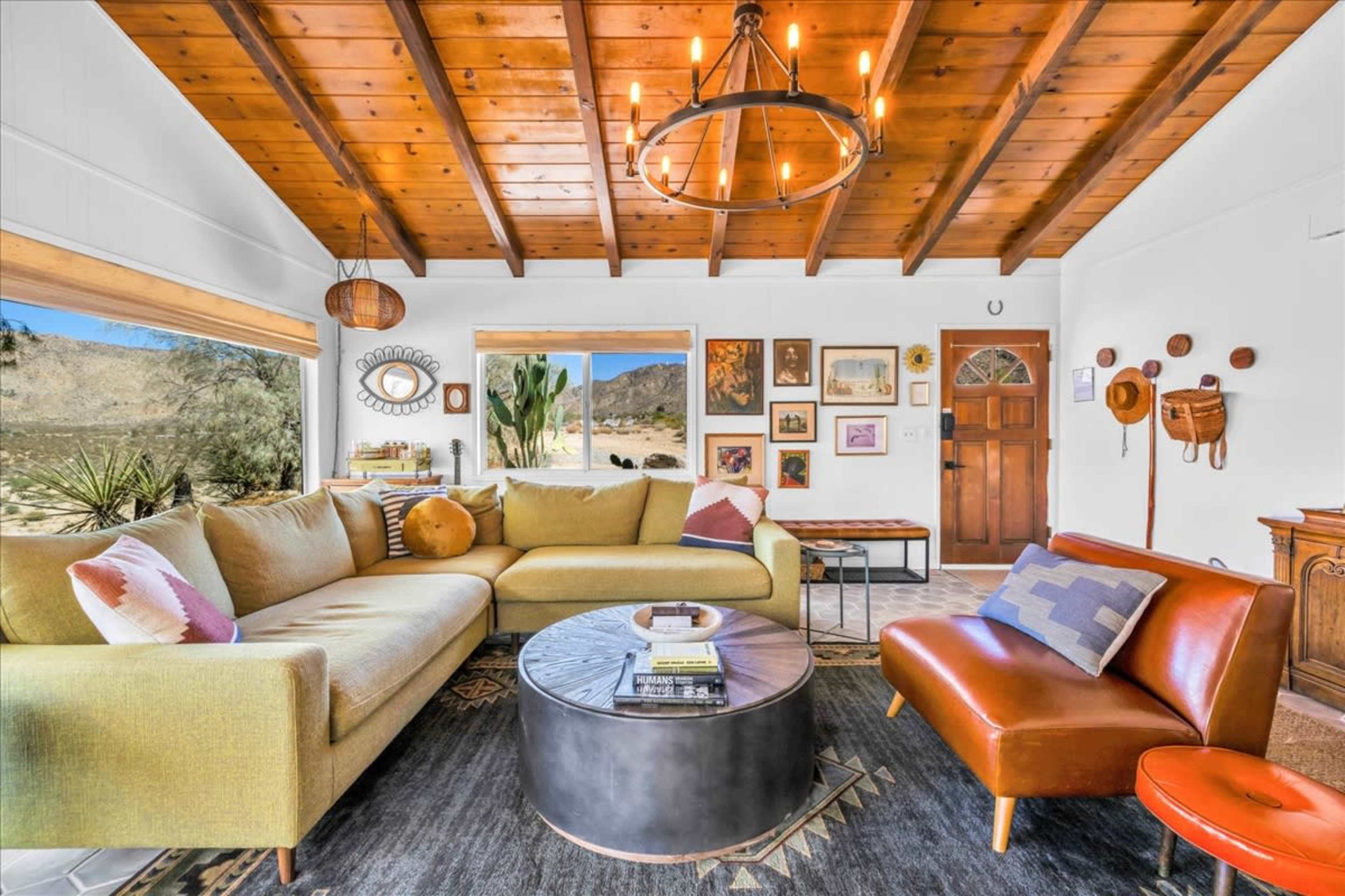 The living room features a large sectional sofa, a round coffee table, and wooden beams on the ceiling, with artwork and decor on the walls and a view of a desert landscape through the window.