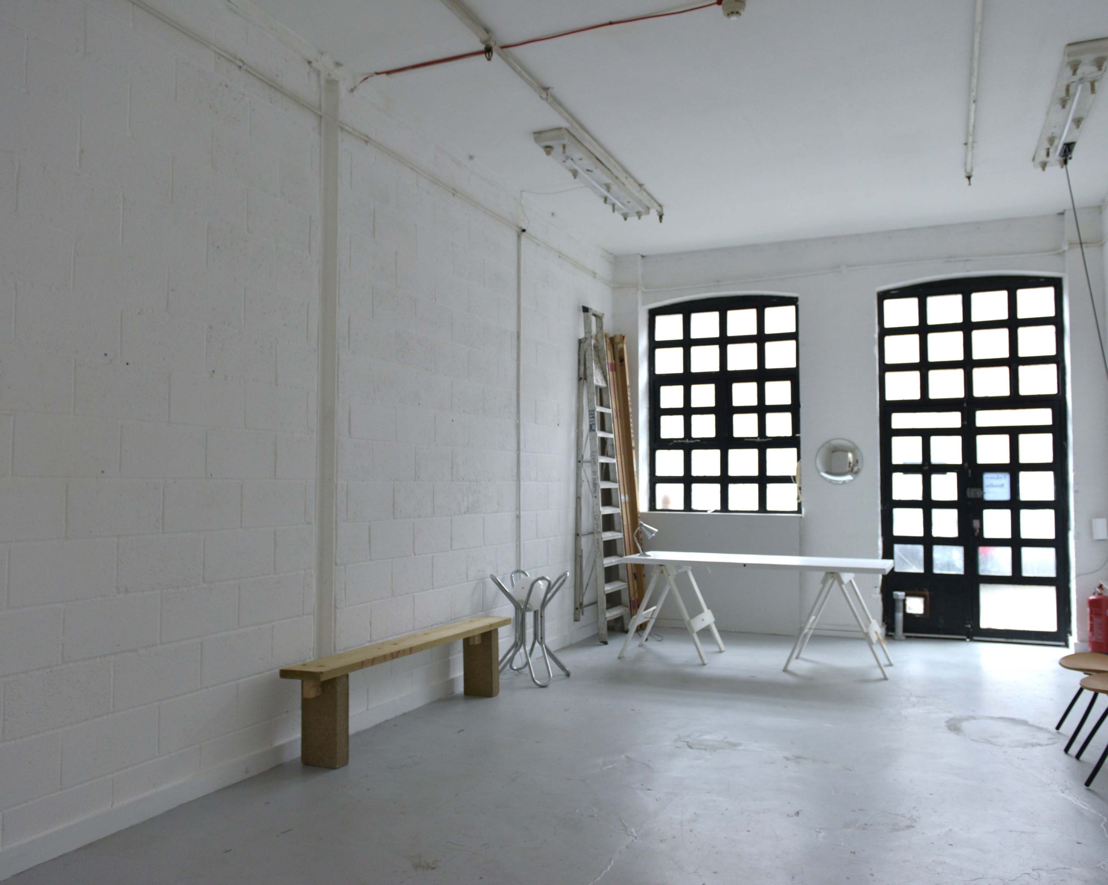 A spacious, empty room with white walls, a wooden bench, a ladder, and a table near large, grid-patterned windows.