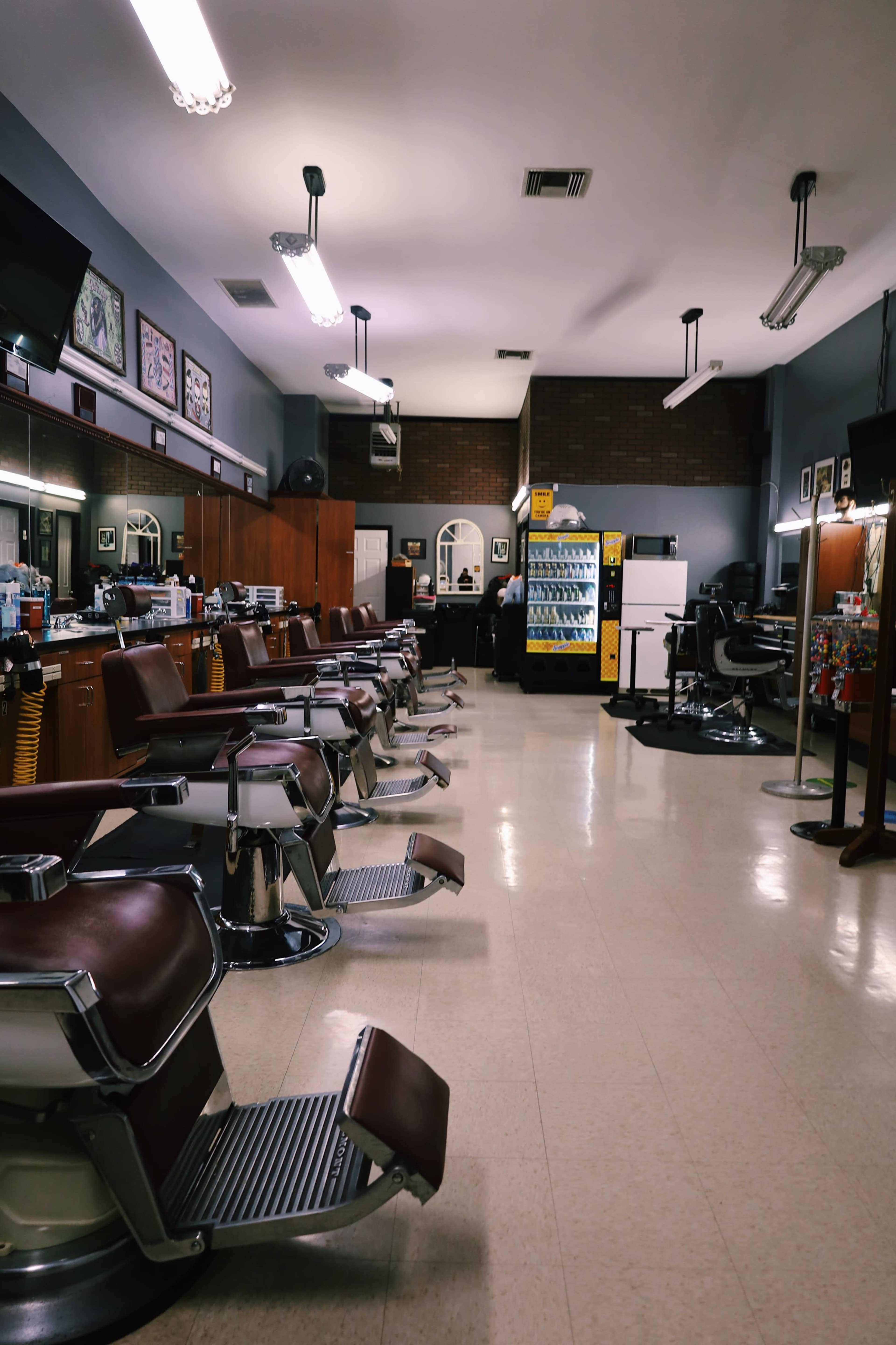 The image shows a row of vintage barber chairs in a spacious barbershop with mirrors, cabinets, and bright overhead lighting.