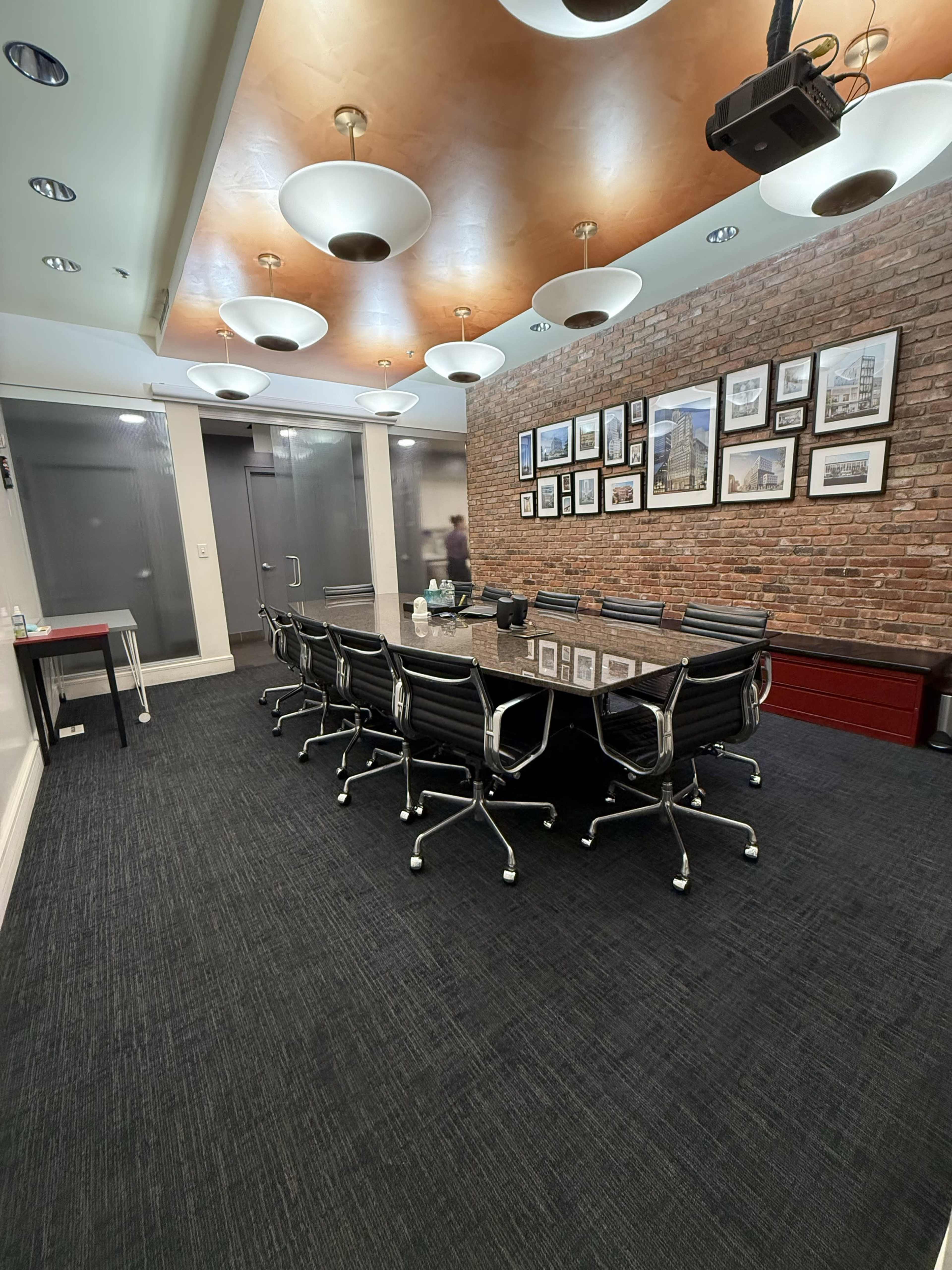 A conference room features a long glass table surrounded by black leather chairs, with exposed brick walls and framed pictures on display.