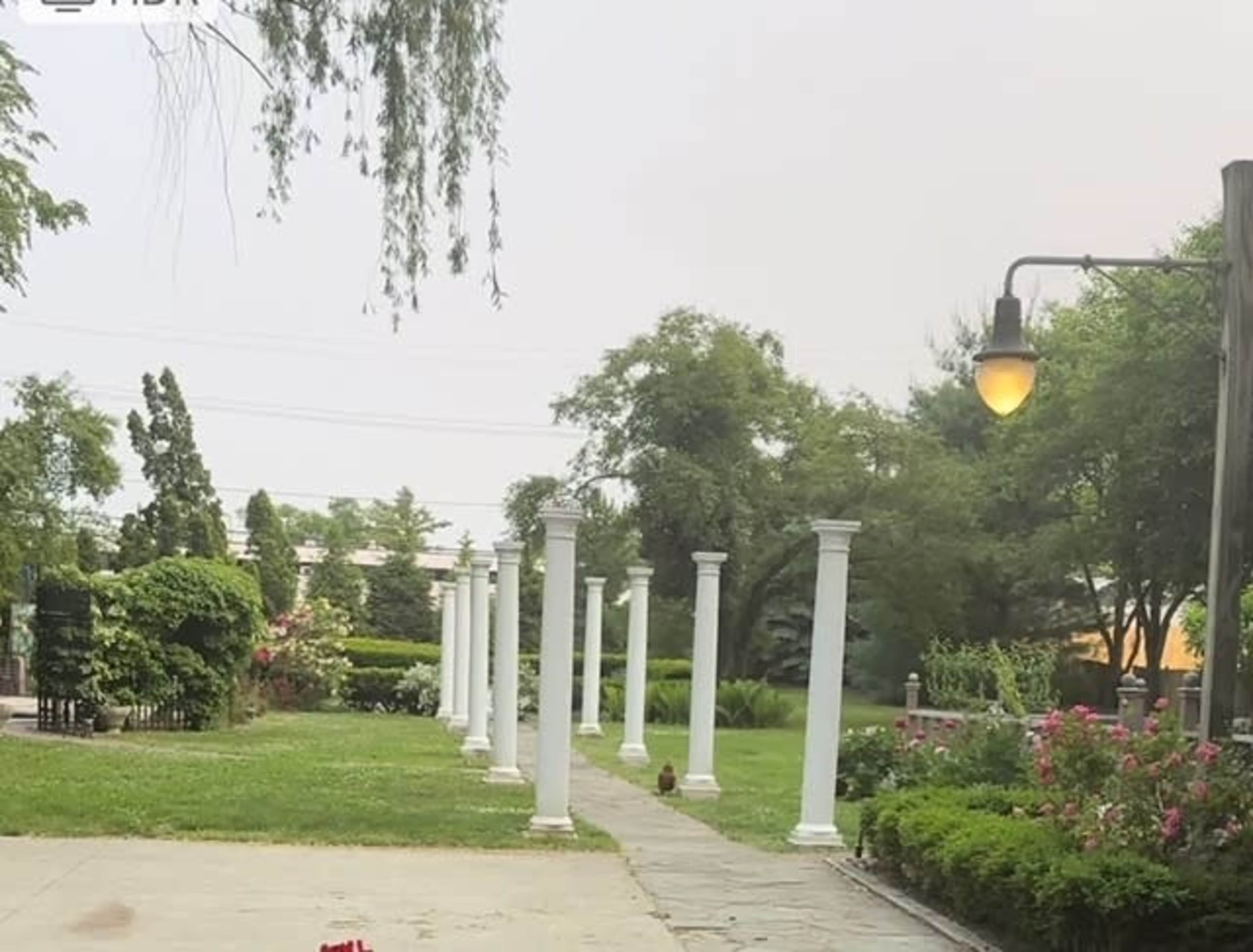 The image shows a walkway lined with white columns leading into a landscaped area with greenery and flowers.