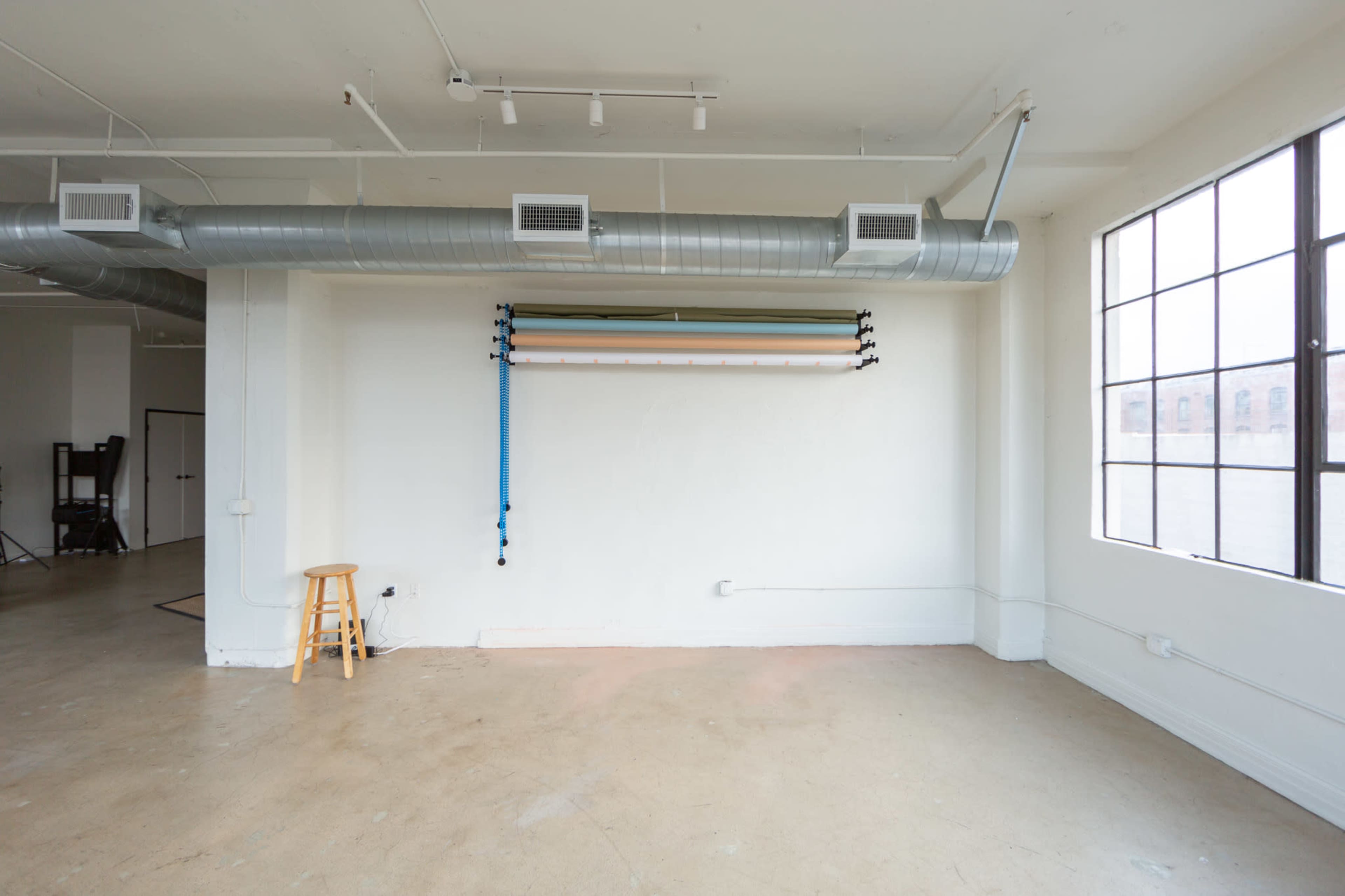 An empty studio space features a white wall with a mounted backdrop system and a wooden stool.