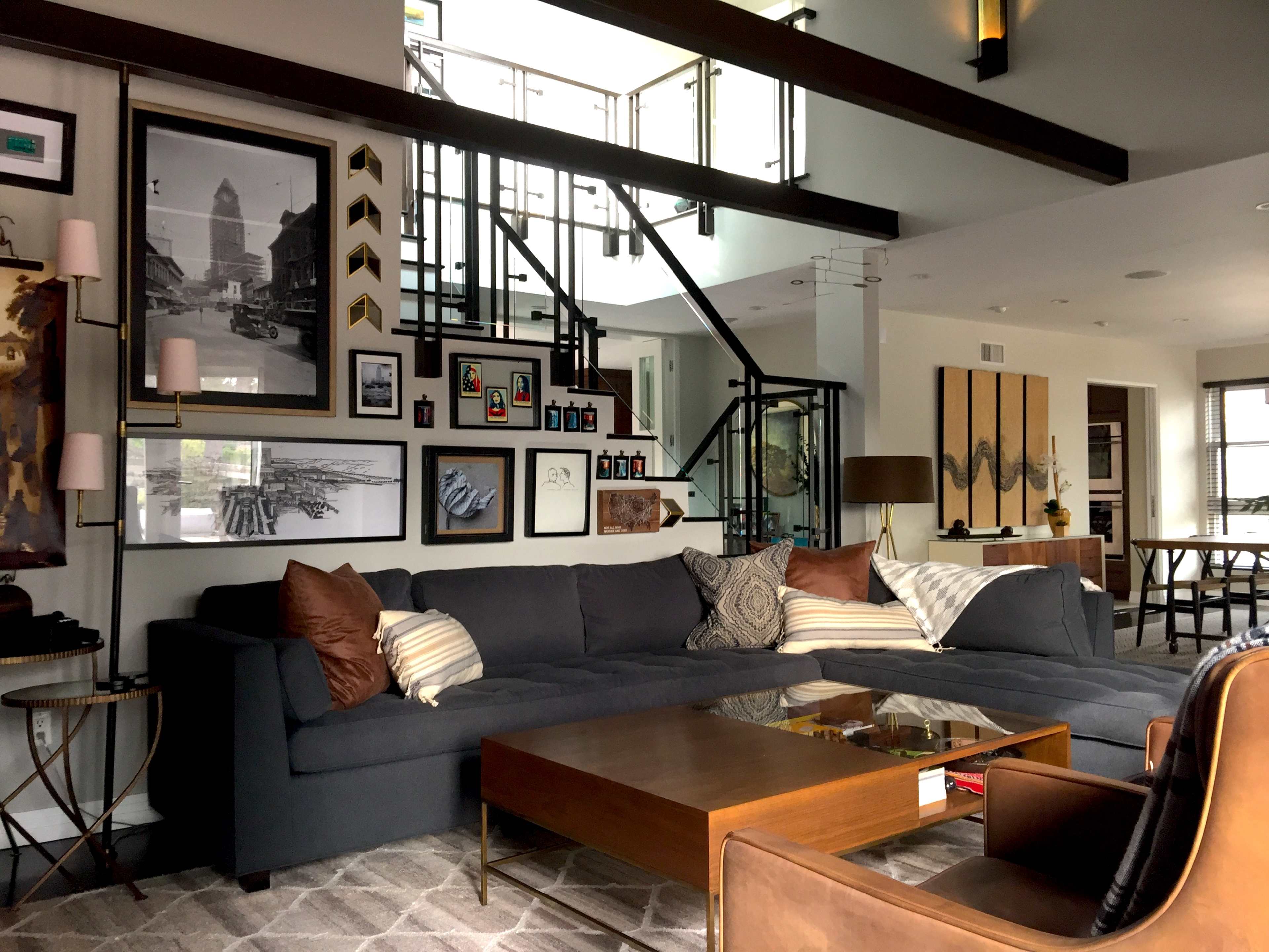 The image shows a modern living room with a large sectional sofa, a wooden coffee table, and a gallery wall featuring various framed photographs and artworks, all set within a contemporary architectural space.