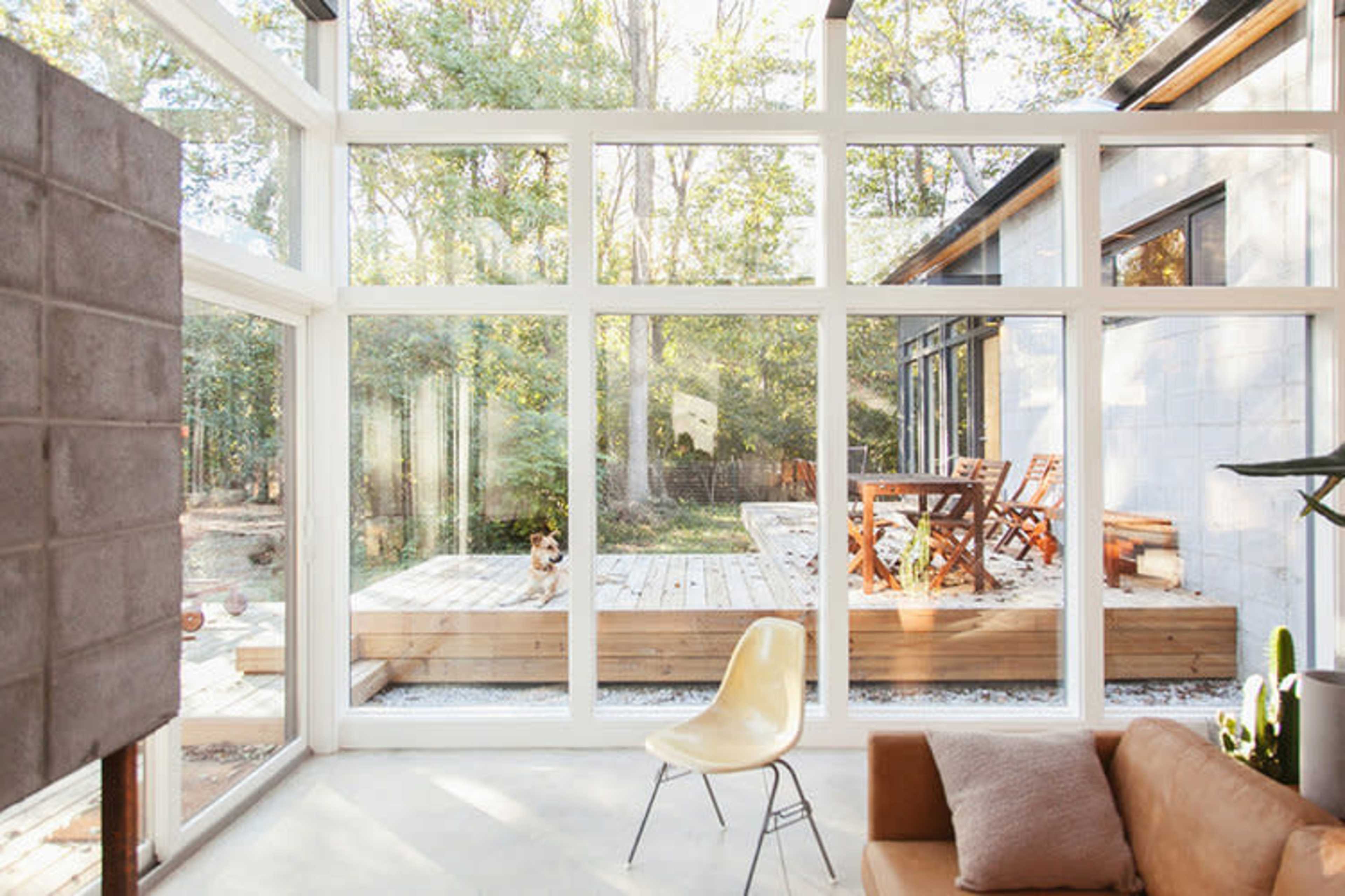 A sunlit living space with large glass windows overlooking a wooden deck and outdoor furniture in a wooded area.
