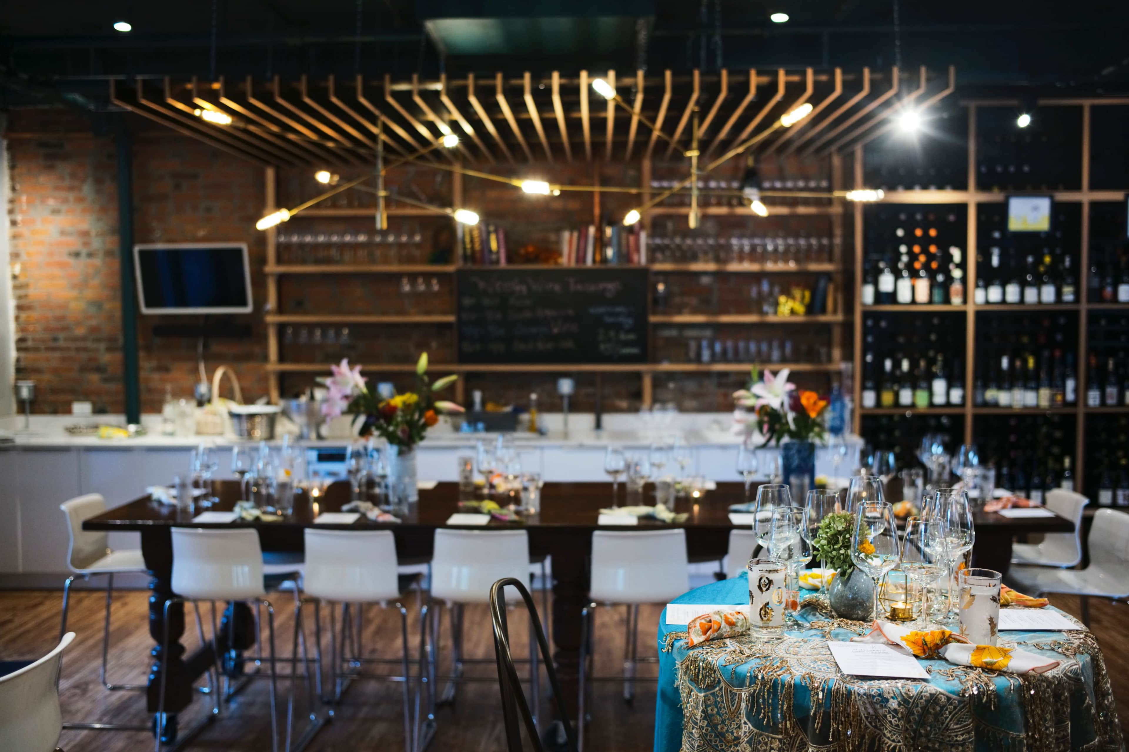 A dining area features a long wooden table set with glassware, surrounded by white chairs, and is decorated with flowers and an assortment of dishes, while a bar area displays shelves of wine.