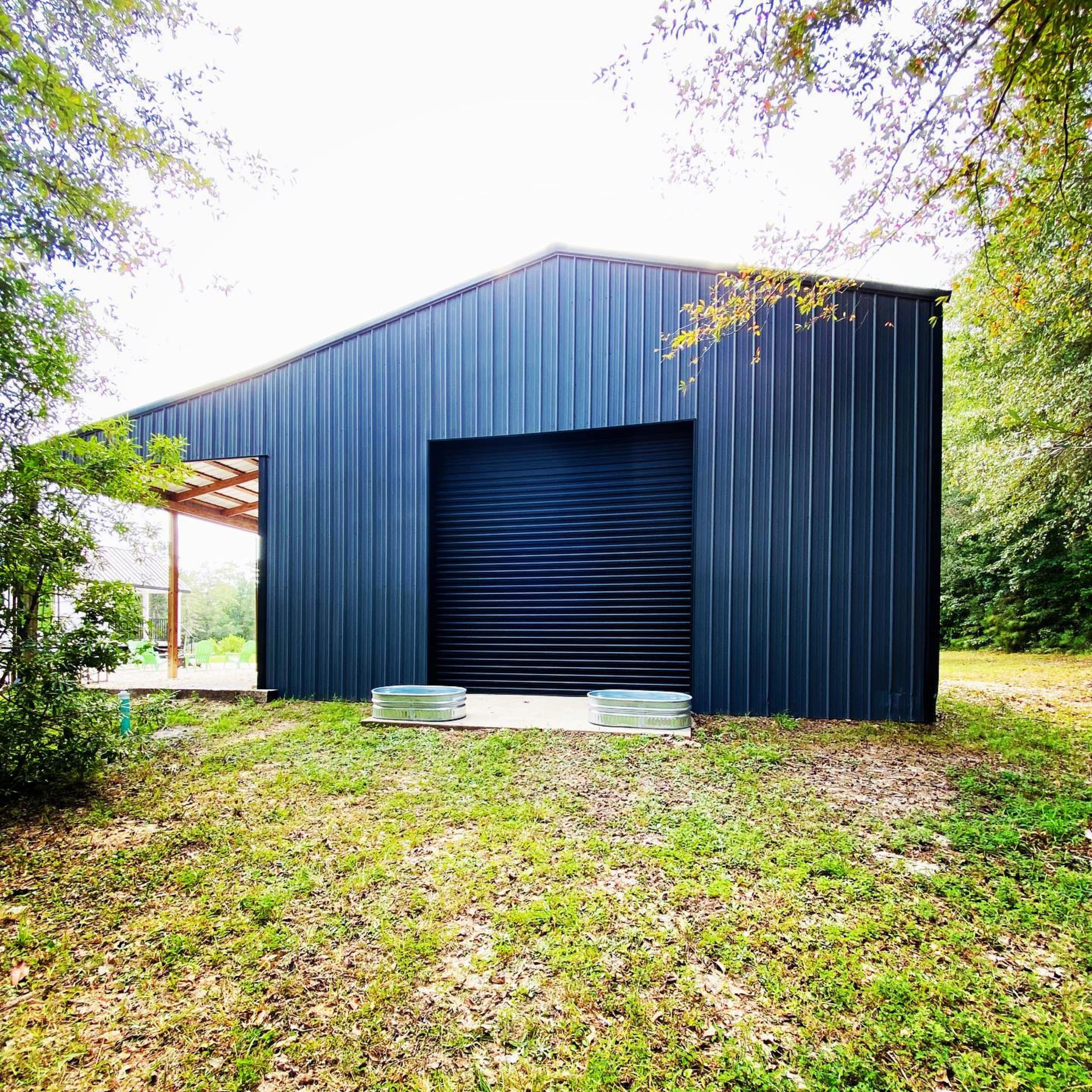 A large, dark metal building with a roll-up door is situated on a grassy area surrounded by trees.