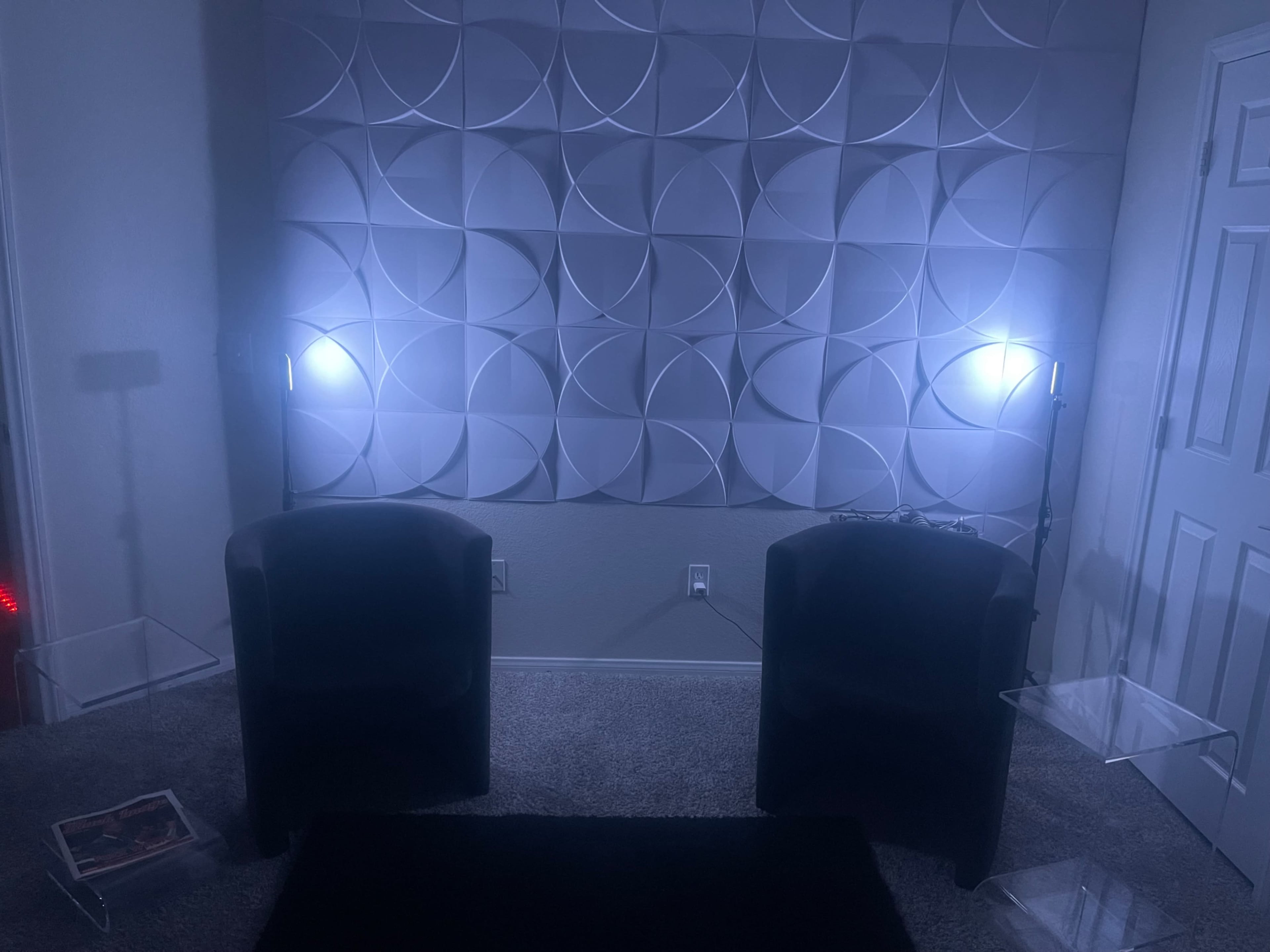 A room with two black chairs facing a textured wall illuminated by soft lighting, with two clear side tables and a dark rug on a carpeted floor.