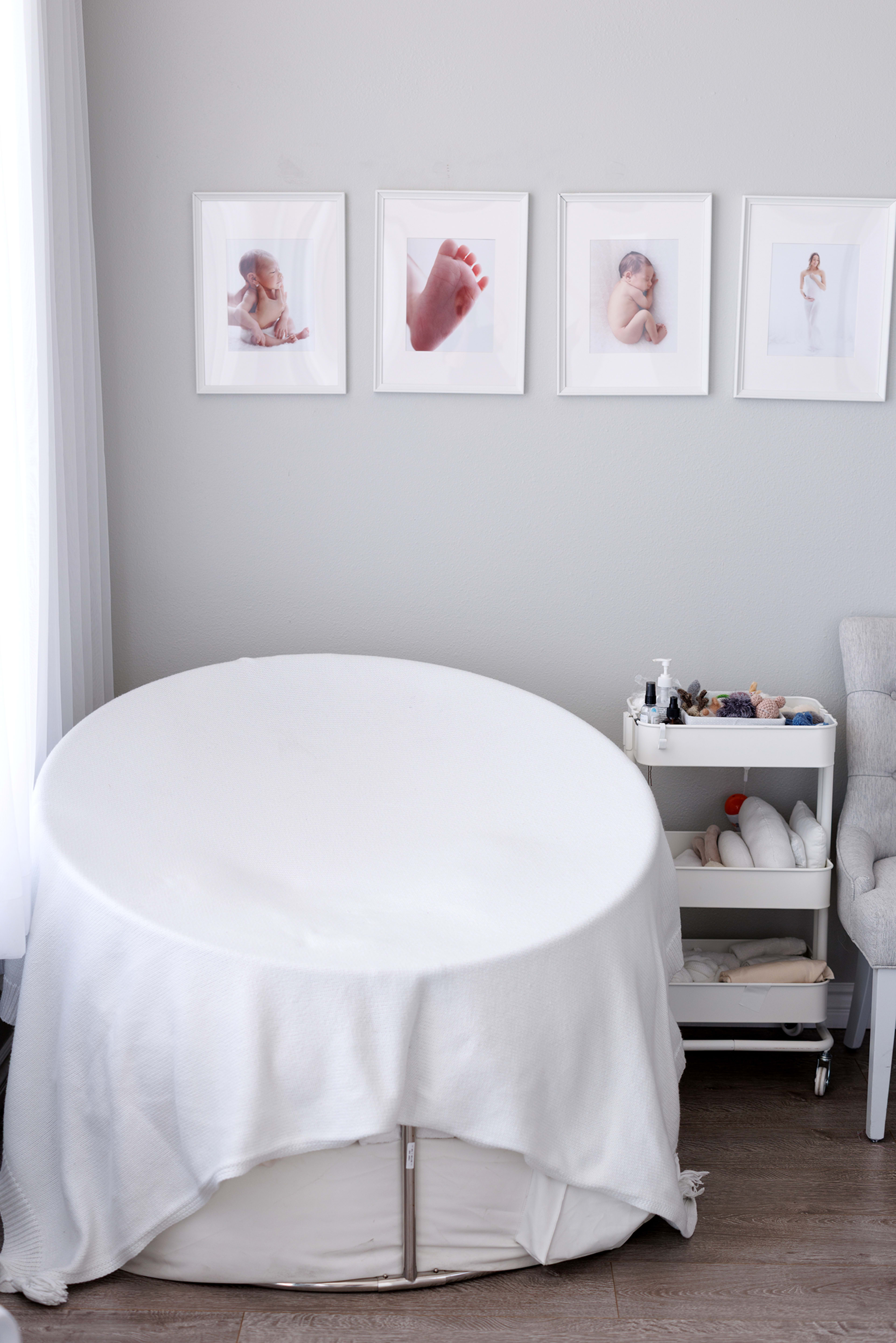 A round massage table, draped with a white cloth, is positioned next to a rolling cart with spa supplies and framed photographs on the wall above.