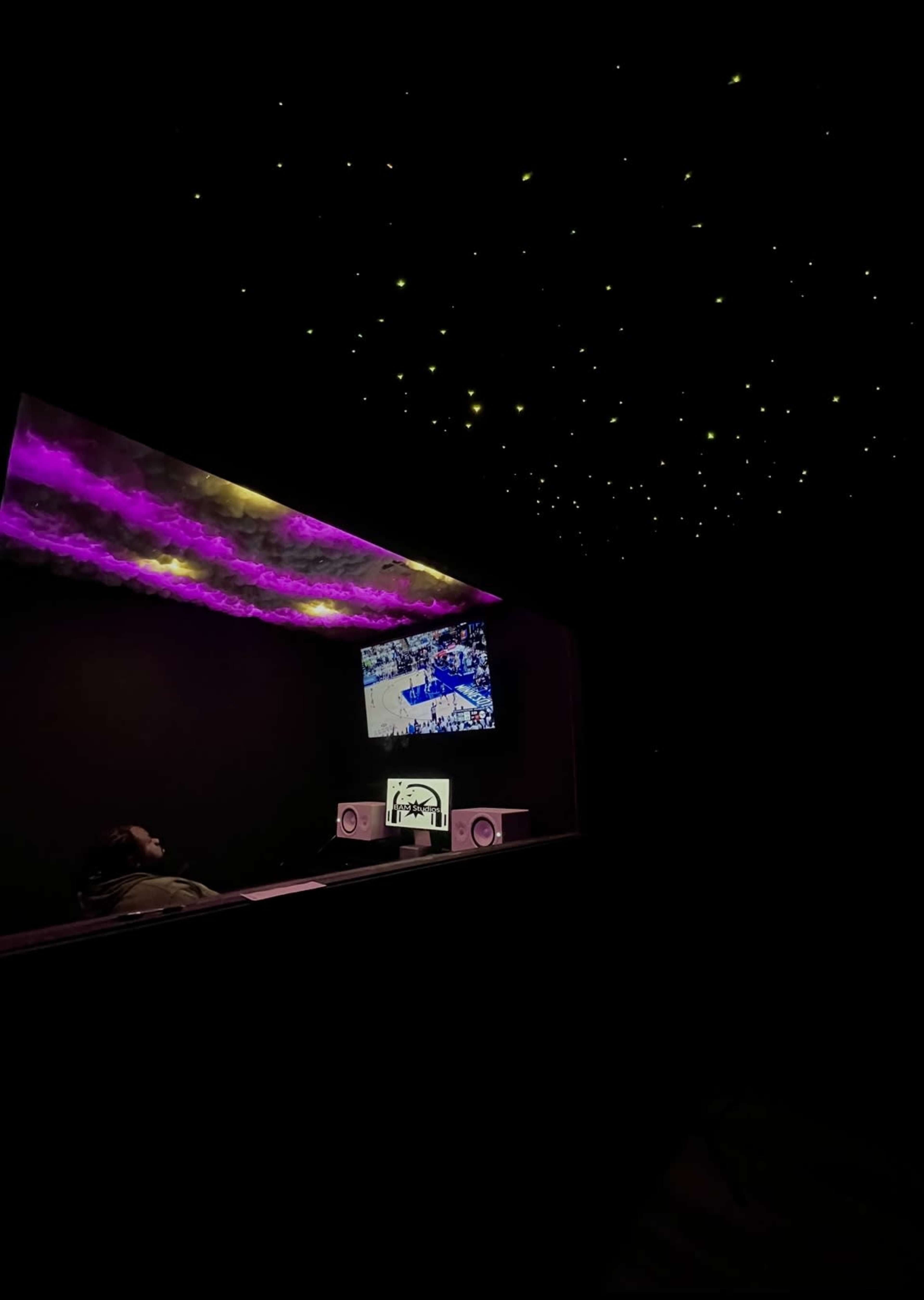 A person sits in a dimly lit room with a monitor displaying a video, while colorful lights illuminated the ceiling.