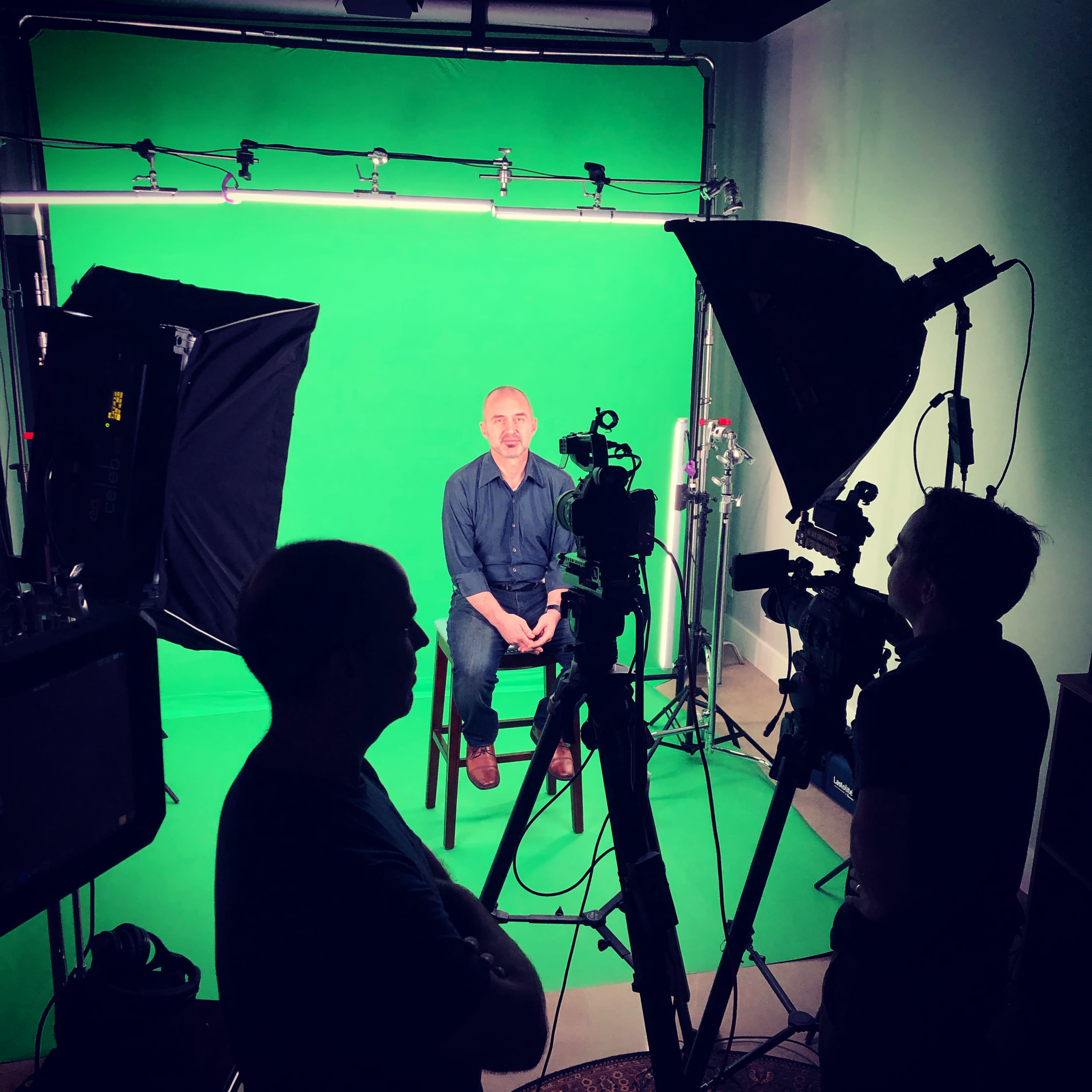 A person sits on a chair in front of a green screen, surrounded by multiple cameras and lighting equipment operated by two crew members.