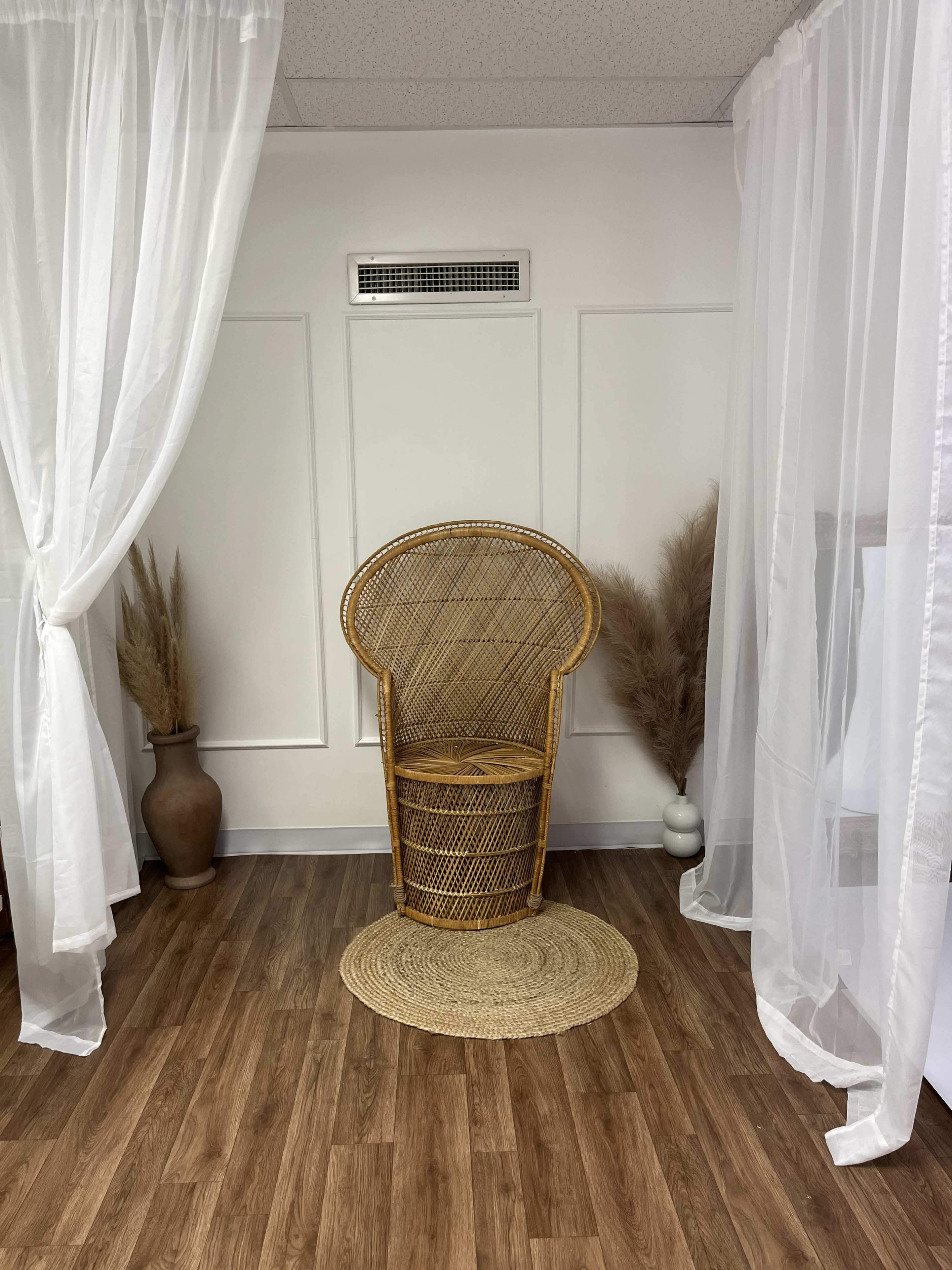 The image shows a well-lit space featuring a rattan chair placed on a round natural fiber rug, surrounded by sheer white curtains and decorative tall grasses in vases.