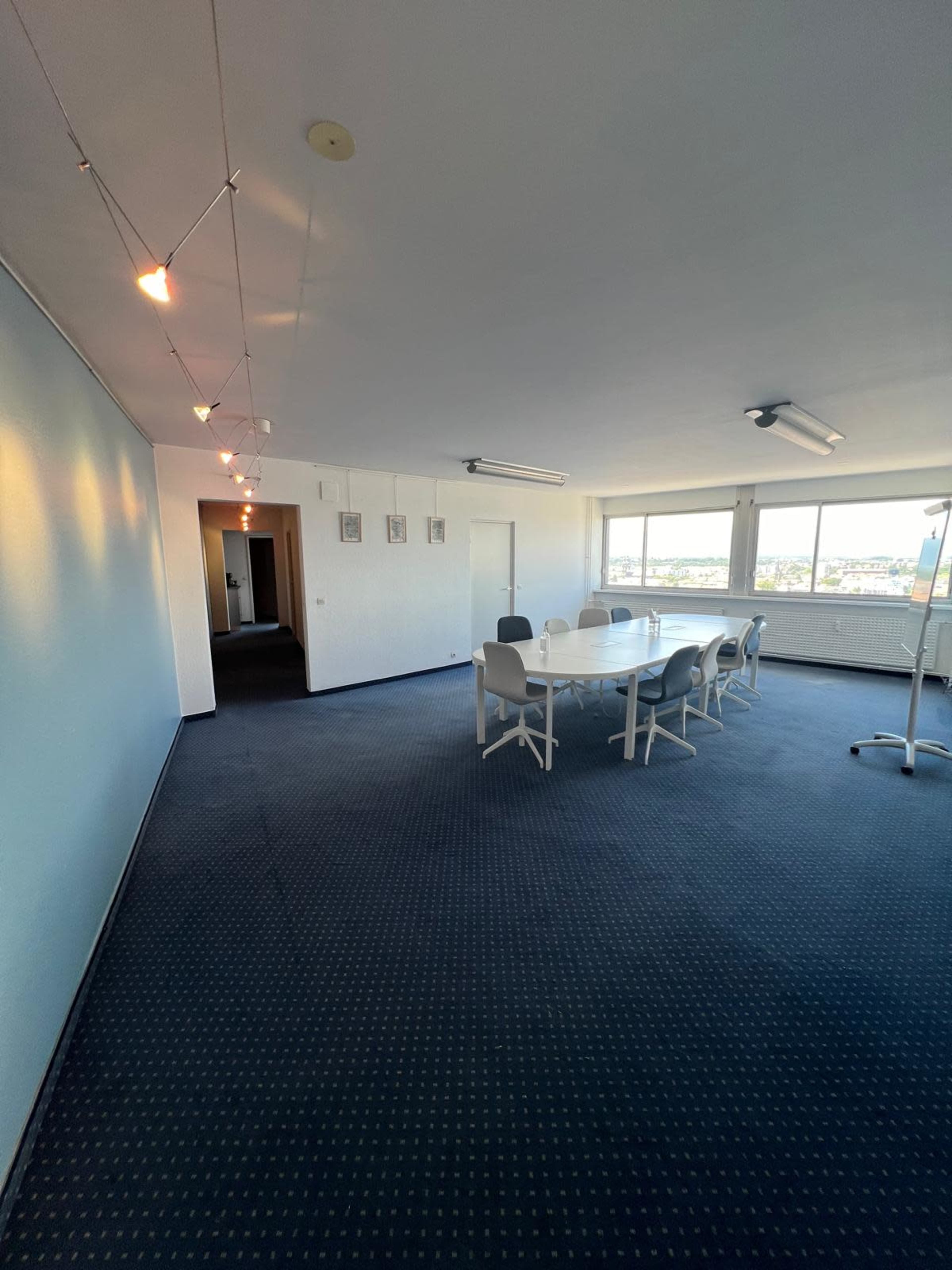 A large meeting room features a circular table surrounded by chairs, with large windows providing natural light and a carpeted floor.