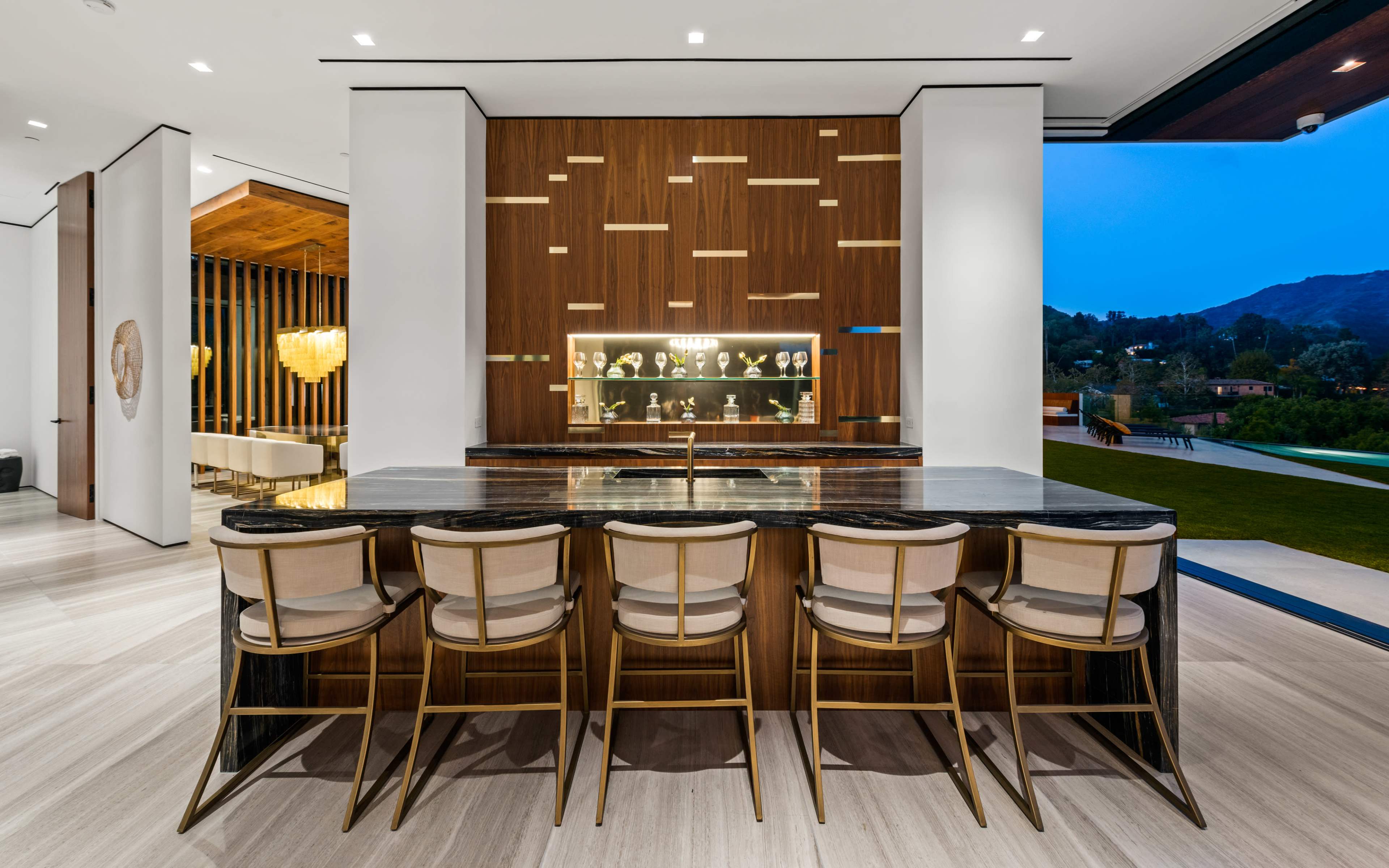 The image shows a modern kitchen bar with a marble countertop and high-backed chairs, set against a wooden accent wall featuring illuminated shelves for displaying items.