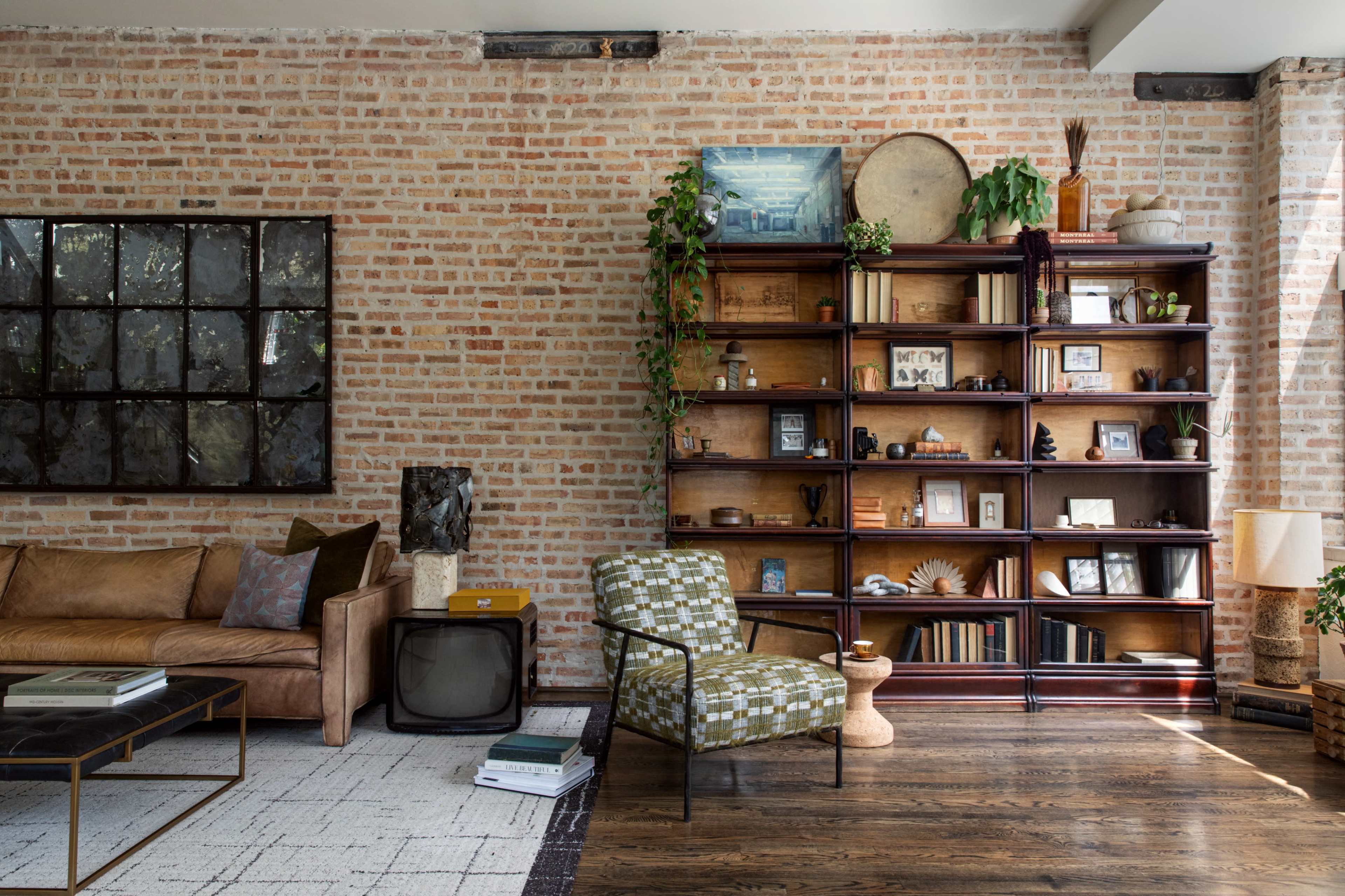 The image shows a living room with a brick wall, a sofa, a patterned chair, a wooden bookshelf filled with various items, and a large window allowing natural light to enter the space.