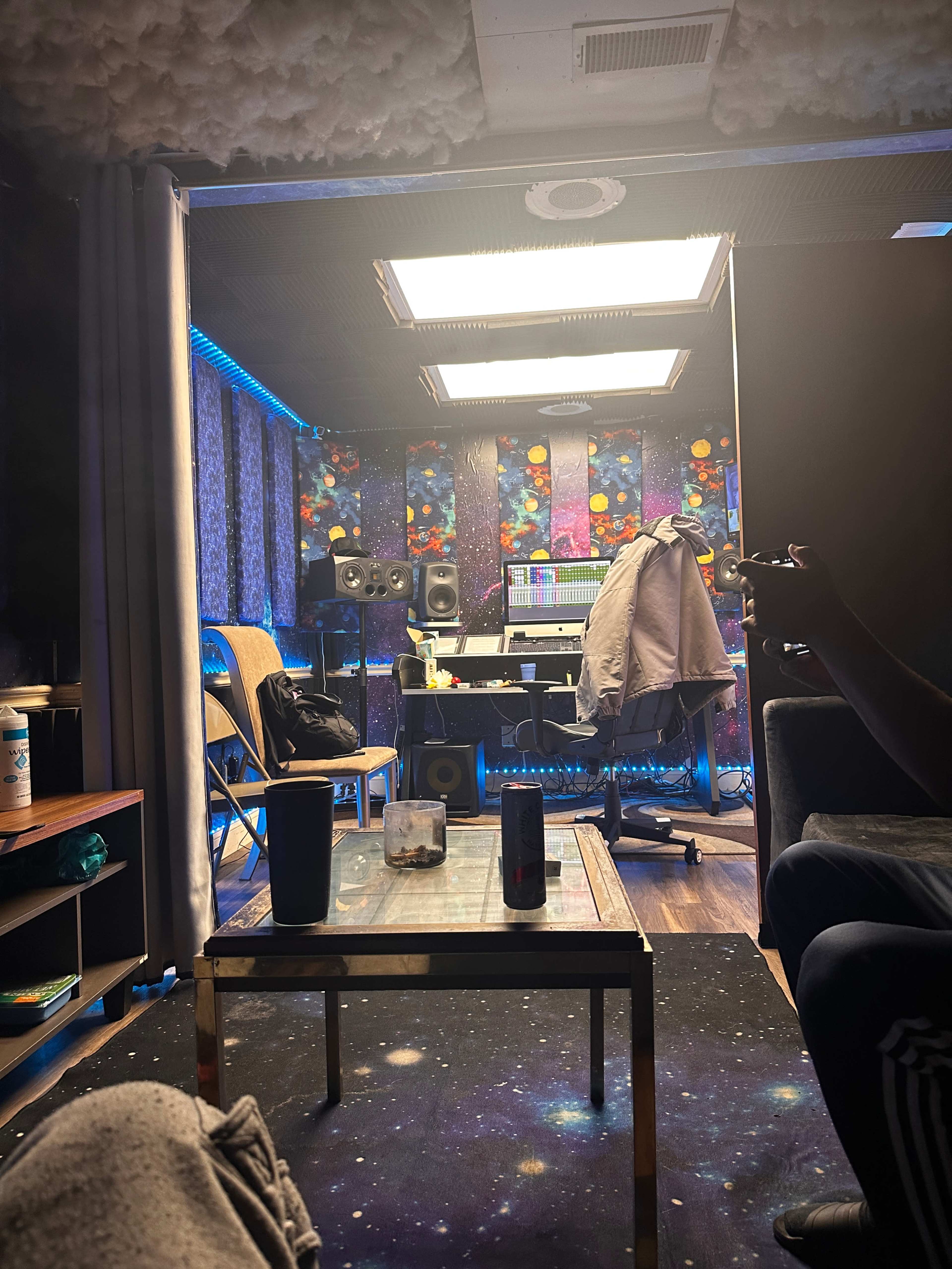 The image shows a cozy music studio with colorful wall art, sound equipment, and a large mixing desk illuminated by blue LED lights.