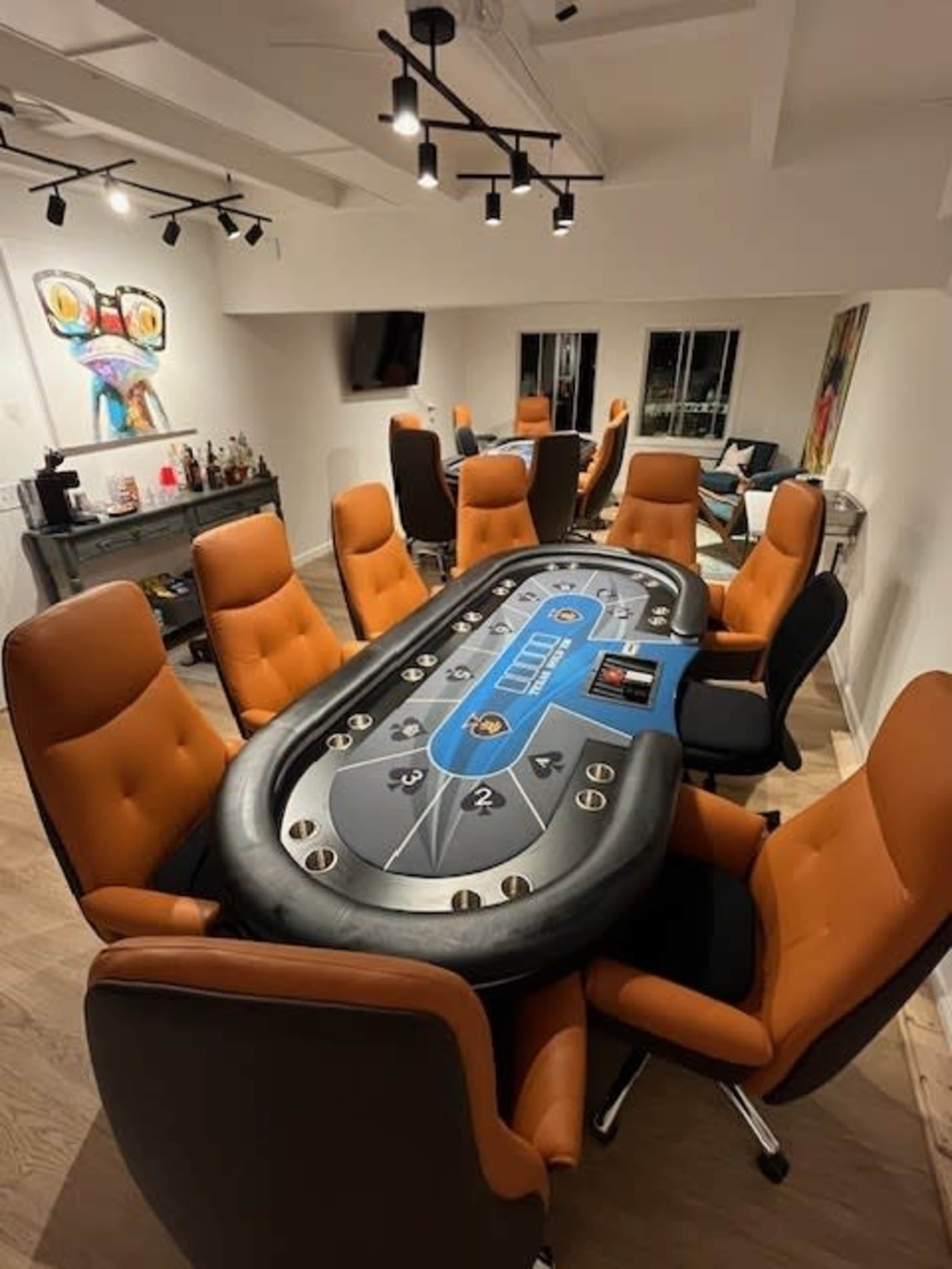 A modern poker room with a large oval table surrounded by orange upholstered chairs and a bar featuring several bottles in the background.