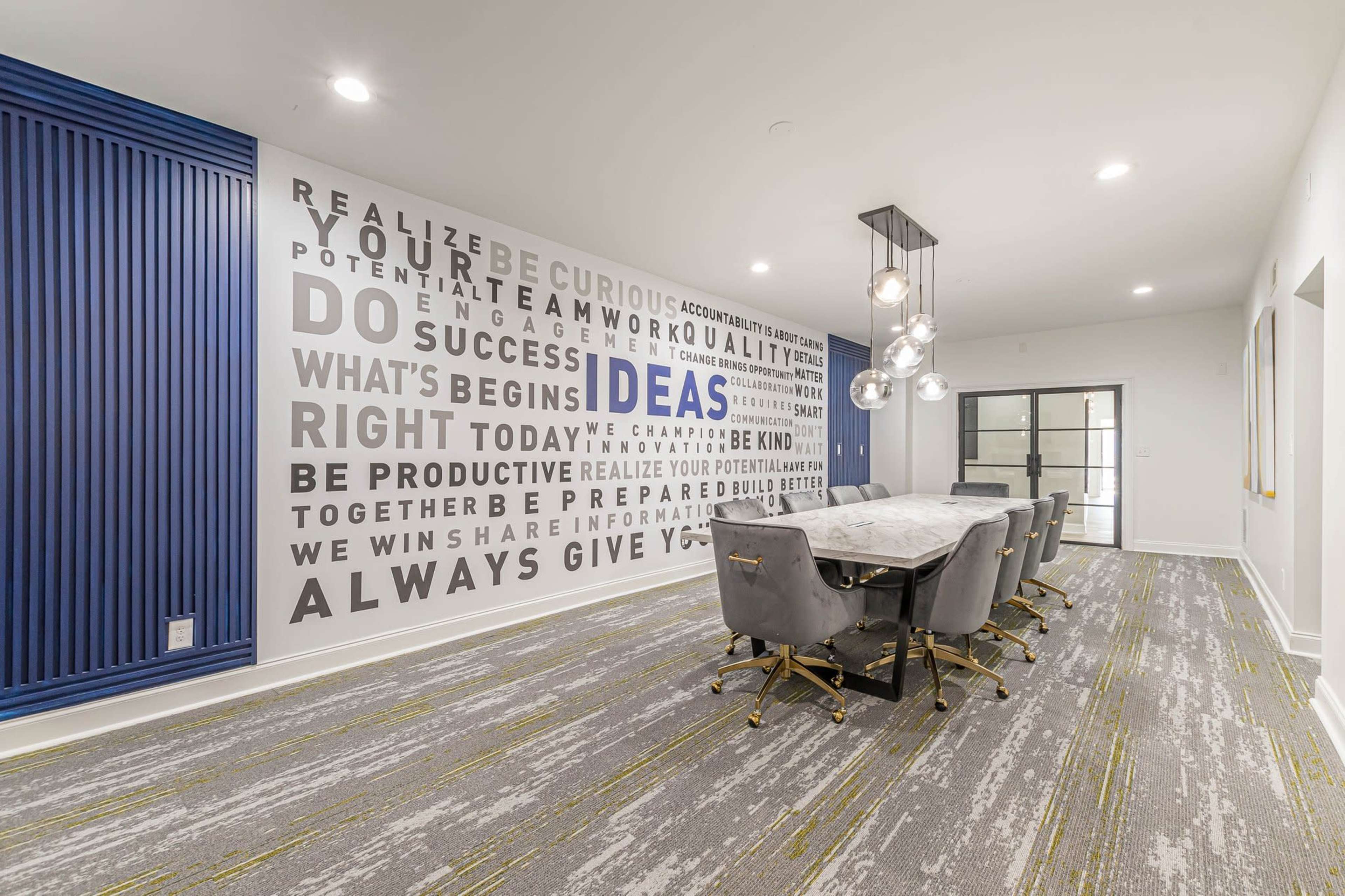 A modern conference room with a large table surrounded by upholstered chairs and a wall featuring motivational text and a pendant light fixture.