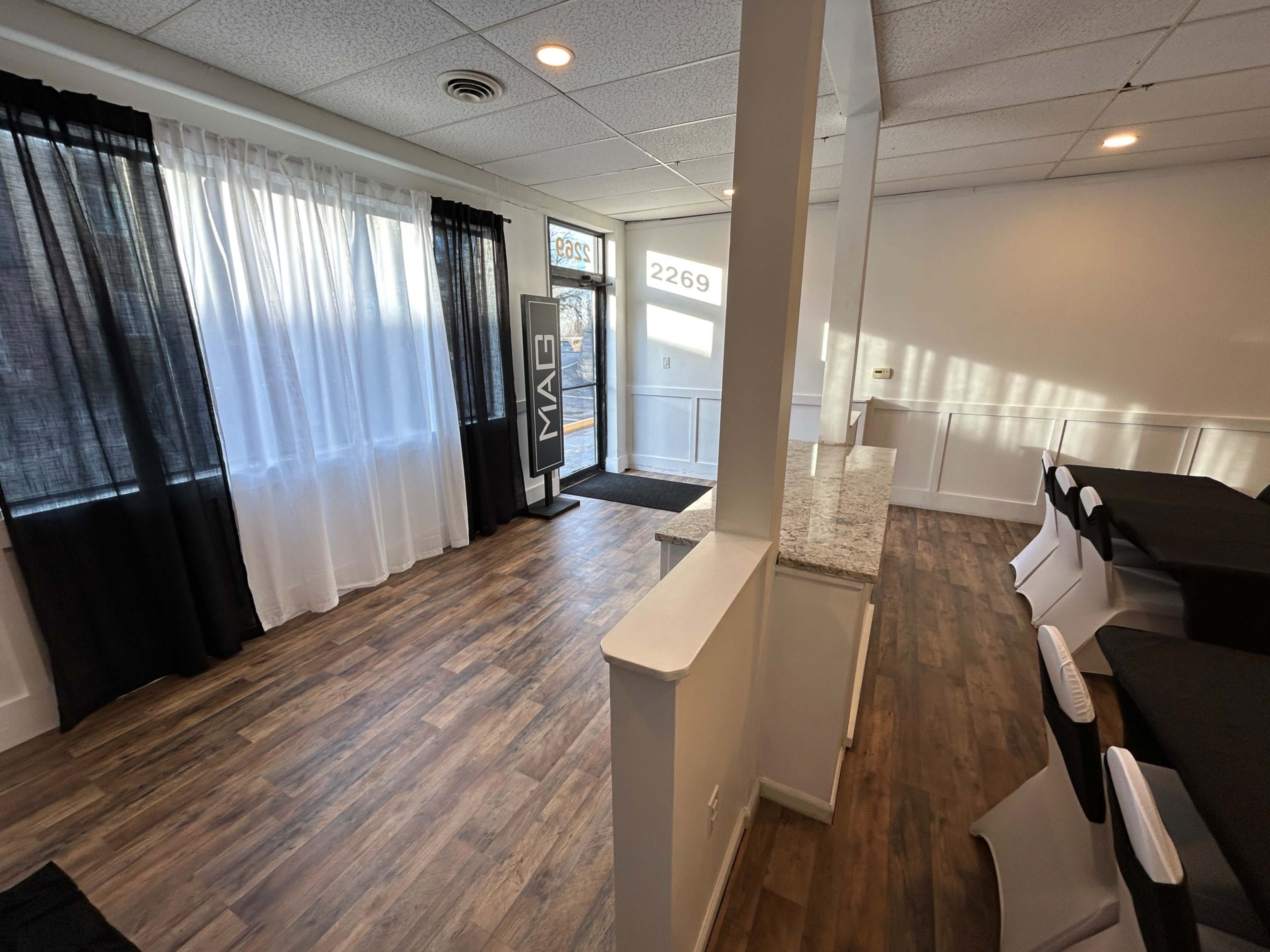 The interior of a bright, empty event space features wooden flooring, black and white decor, and large windows with sheer curtains.
