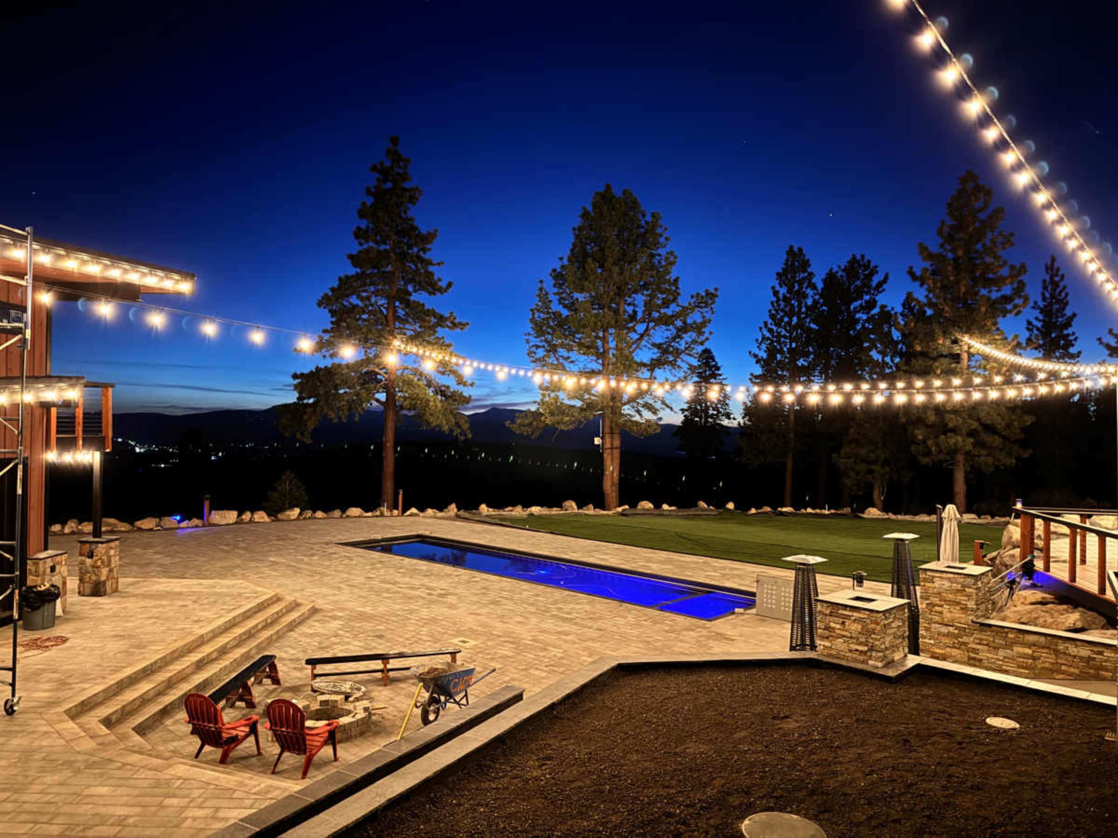 A luxurious outdoor space at night features a pool with blue lights, surrounded by trees and illuminated by string lights.