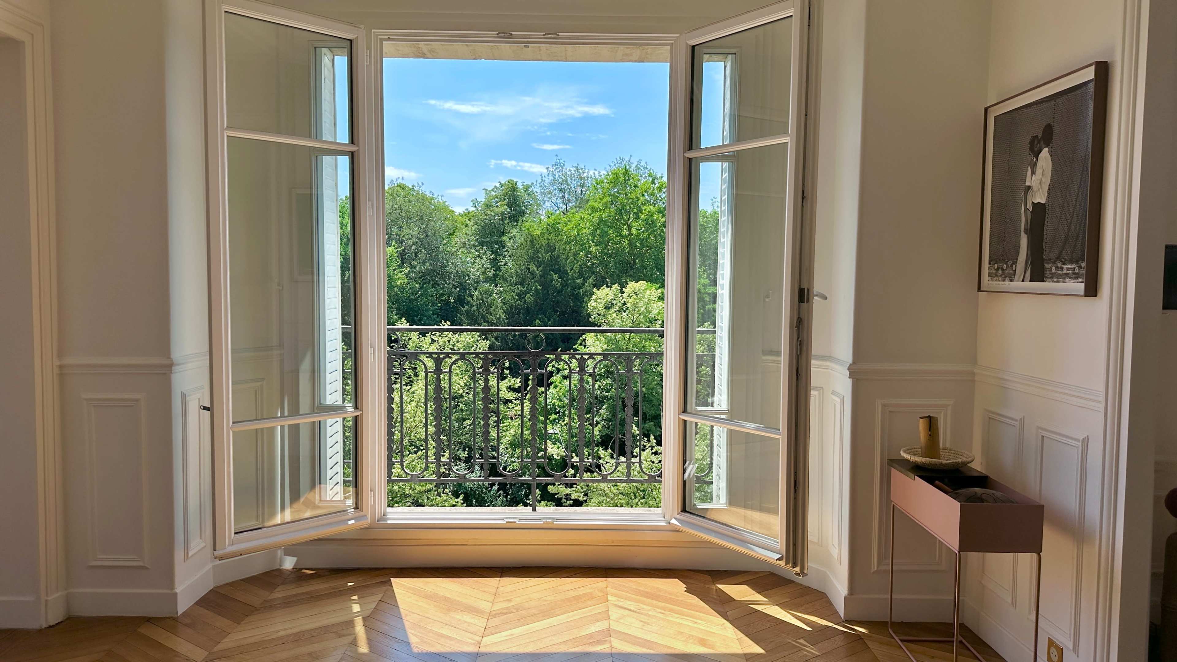 A wide-open window with a wrought-iron balcony overlooks a lush green landscape.
