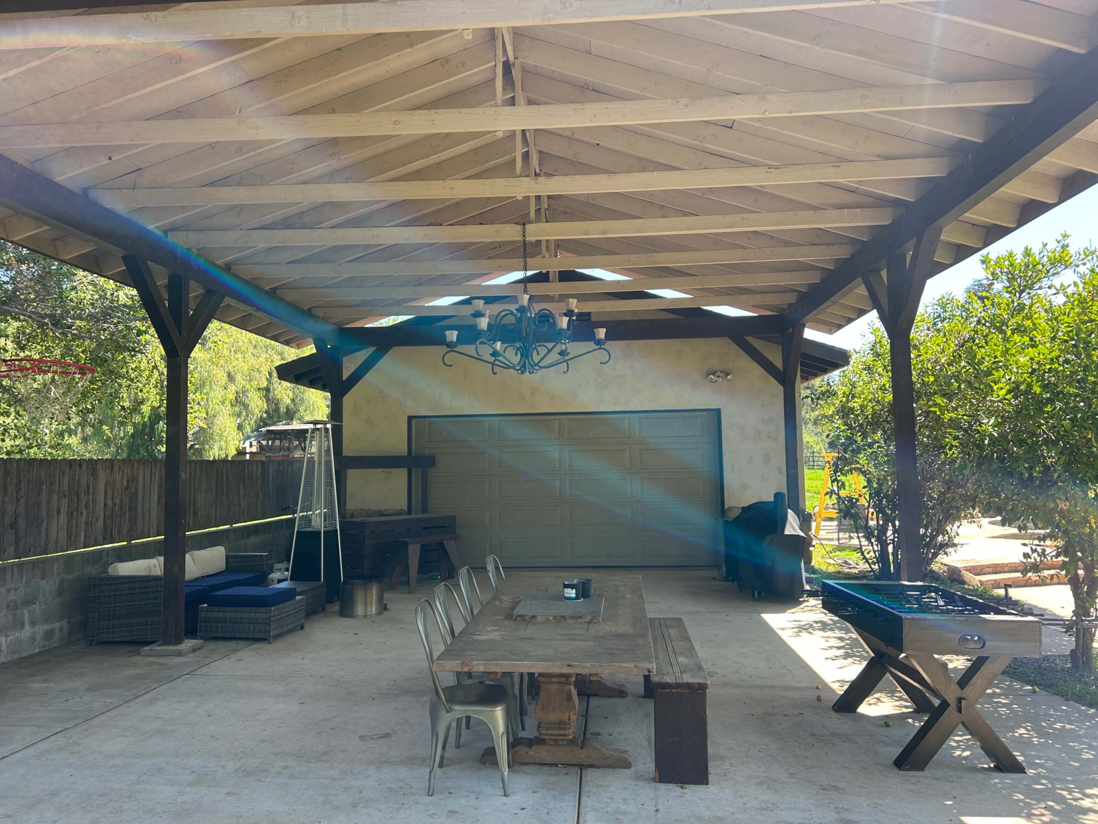 The image shows a covered outdoor patio area with a long wooden table, chairs, and a chandelier, alongside a garage door and landscaping featuring trees.