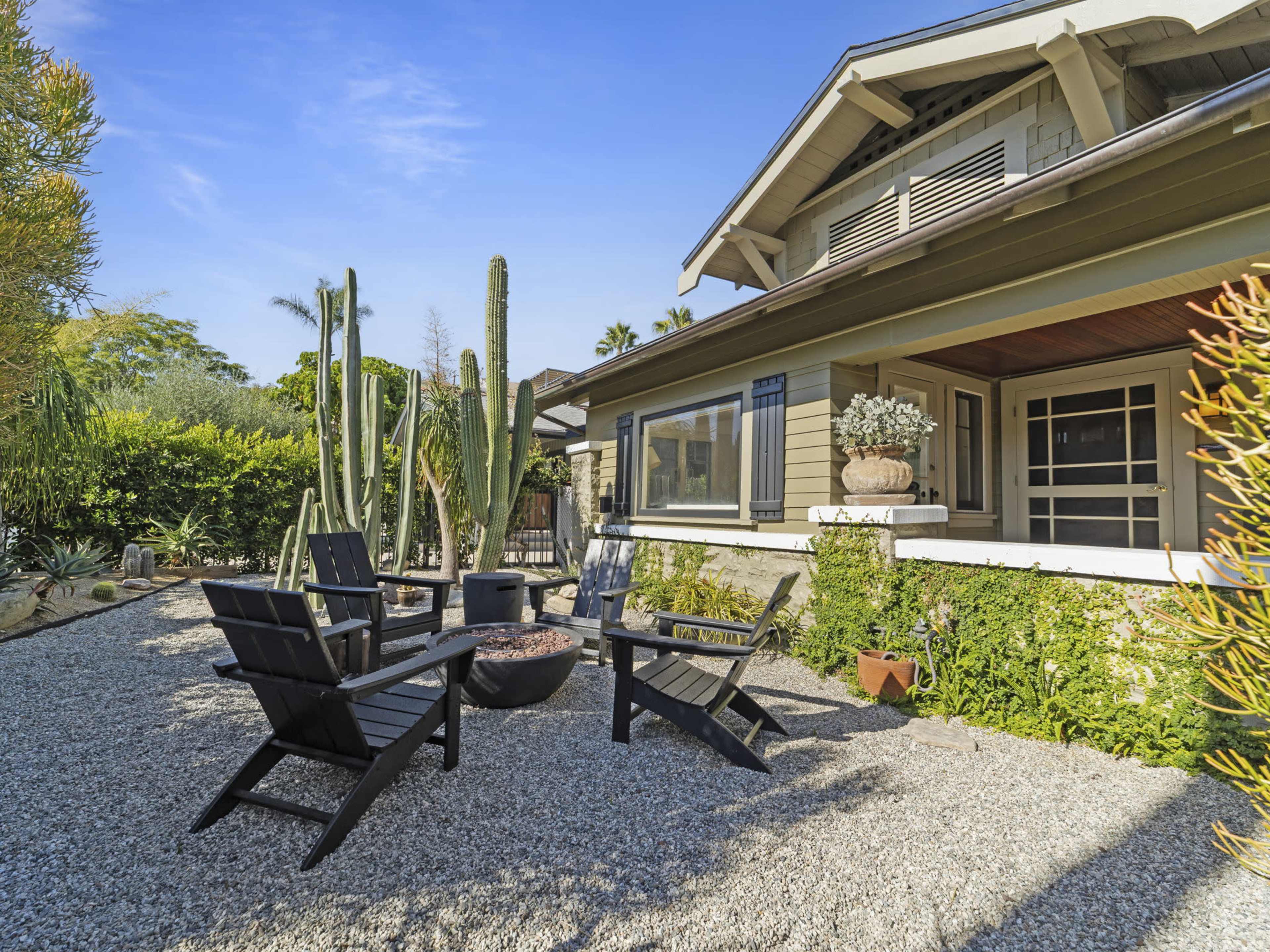 The image shows a landscaped outdoor seating area with four black chairs arranged around a fire pit, surrounded by cacti and greenery near a house.
