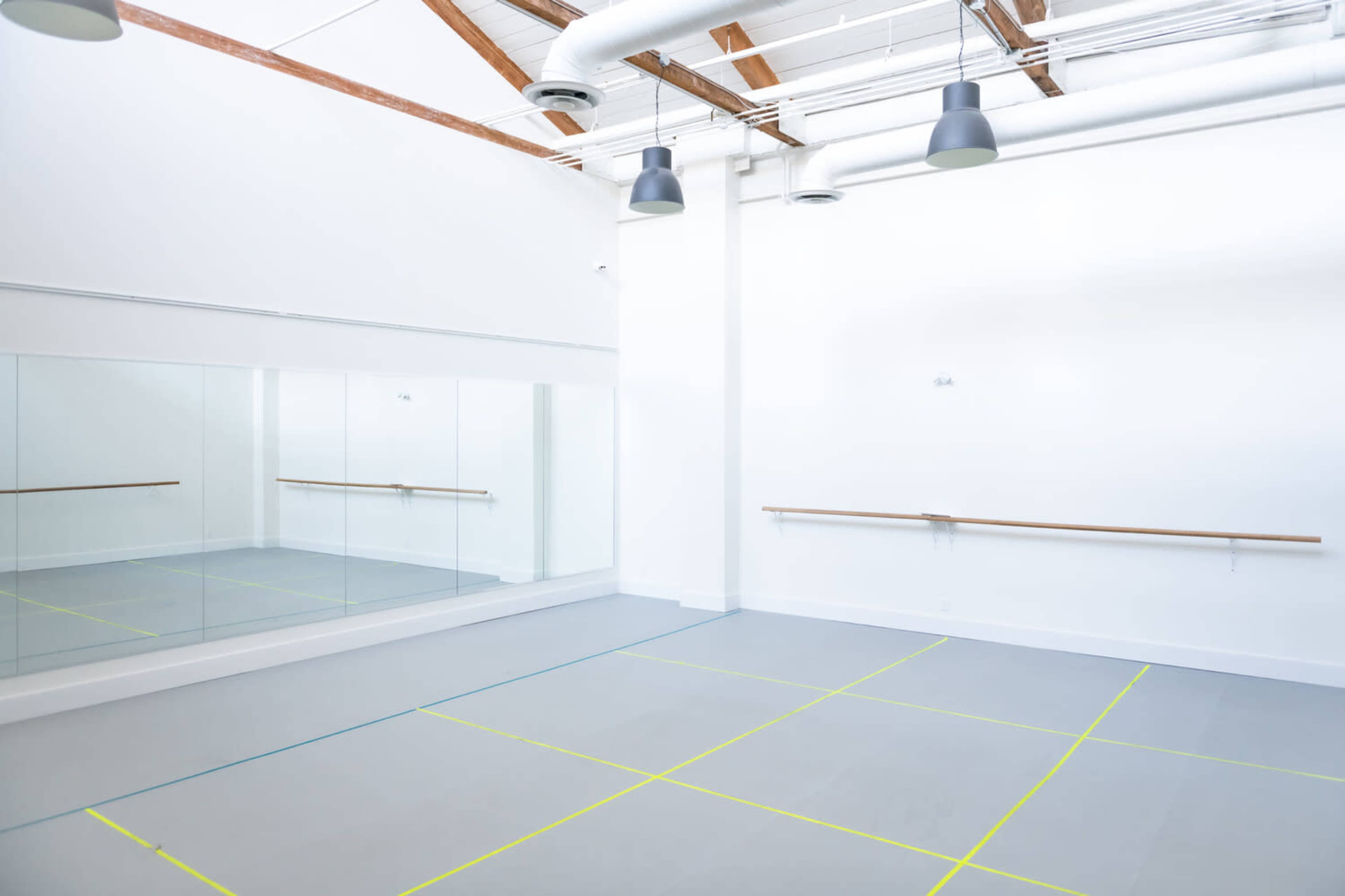 A spacious, brightly lit dance studio featuring mirrors on one wall and a ballet barre along another, with marked floor areas.