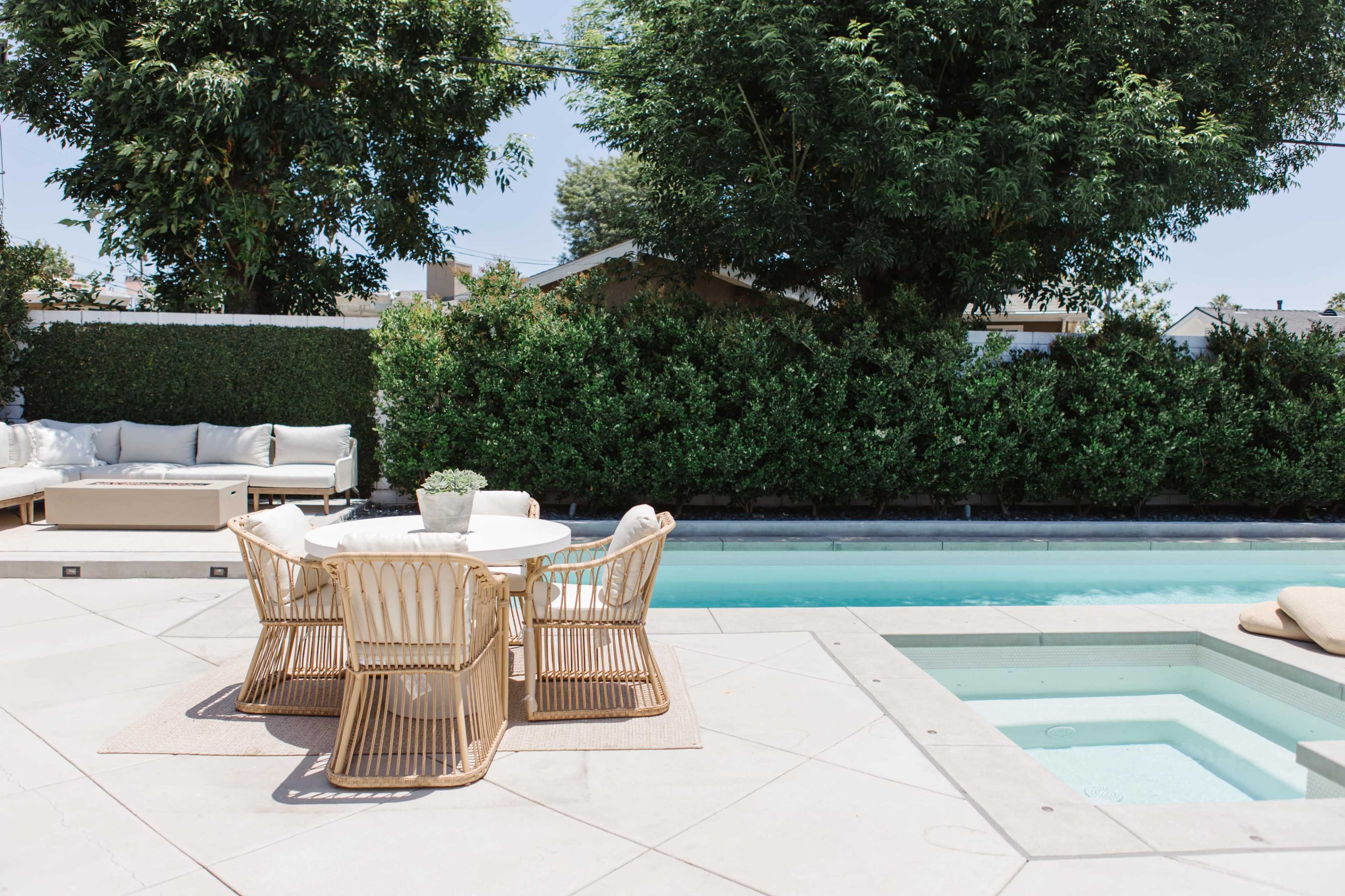 A patio area features a round table with four rattan chairs, adjacent to a swimming pool surrounded by greenery and lounge furniture.