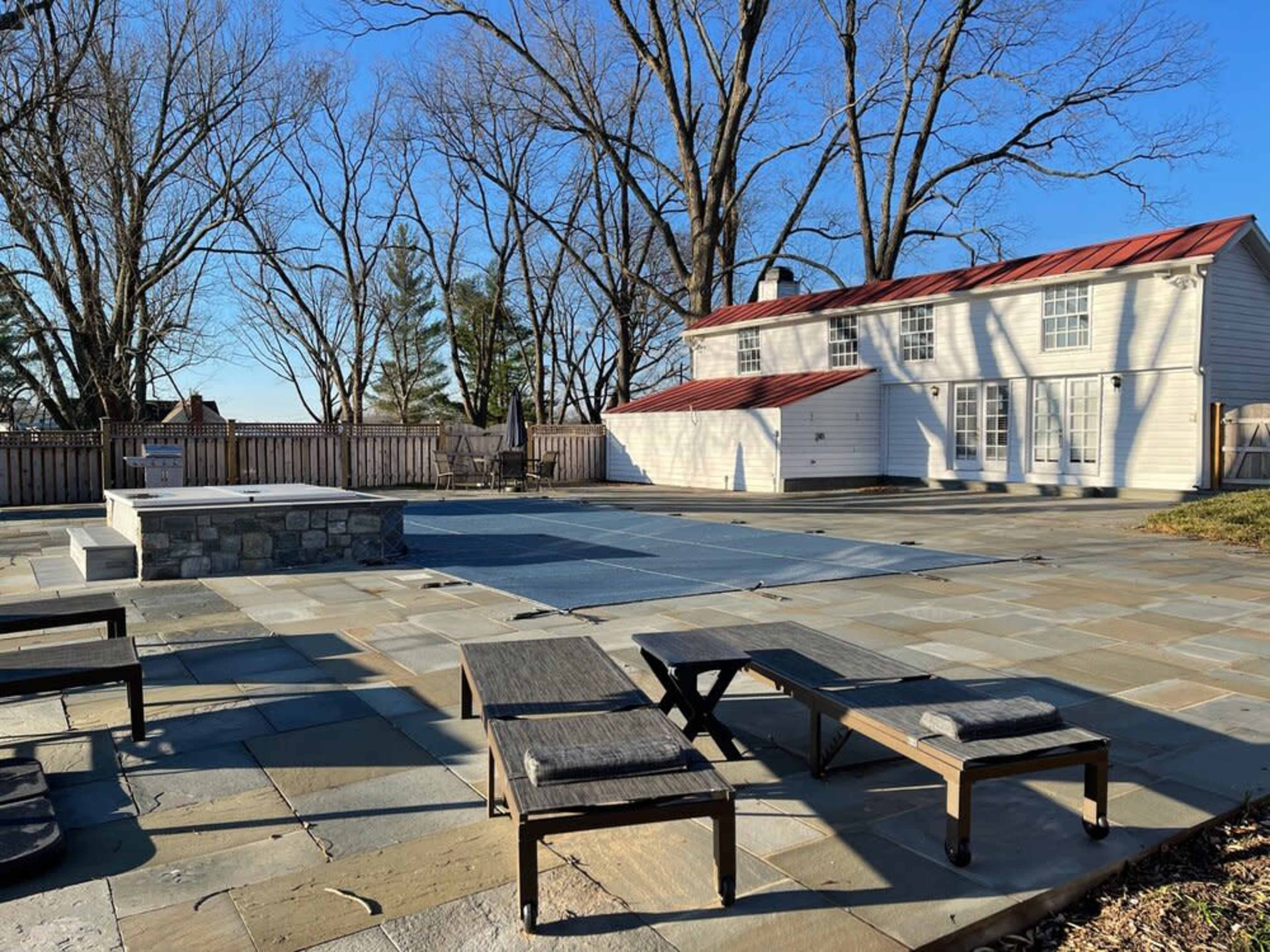 A paved patio area with lounge chairs near a covered swimming pool and a white house with a red roof in the background, surrounded by bare trees.
