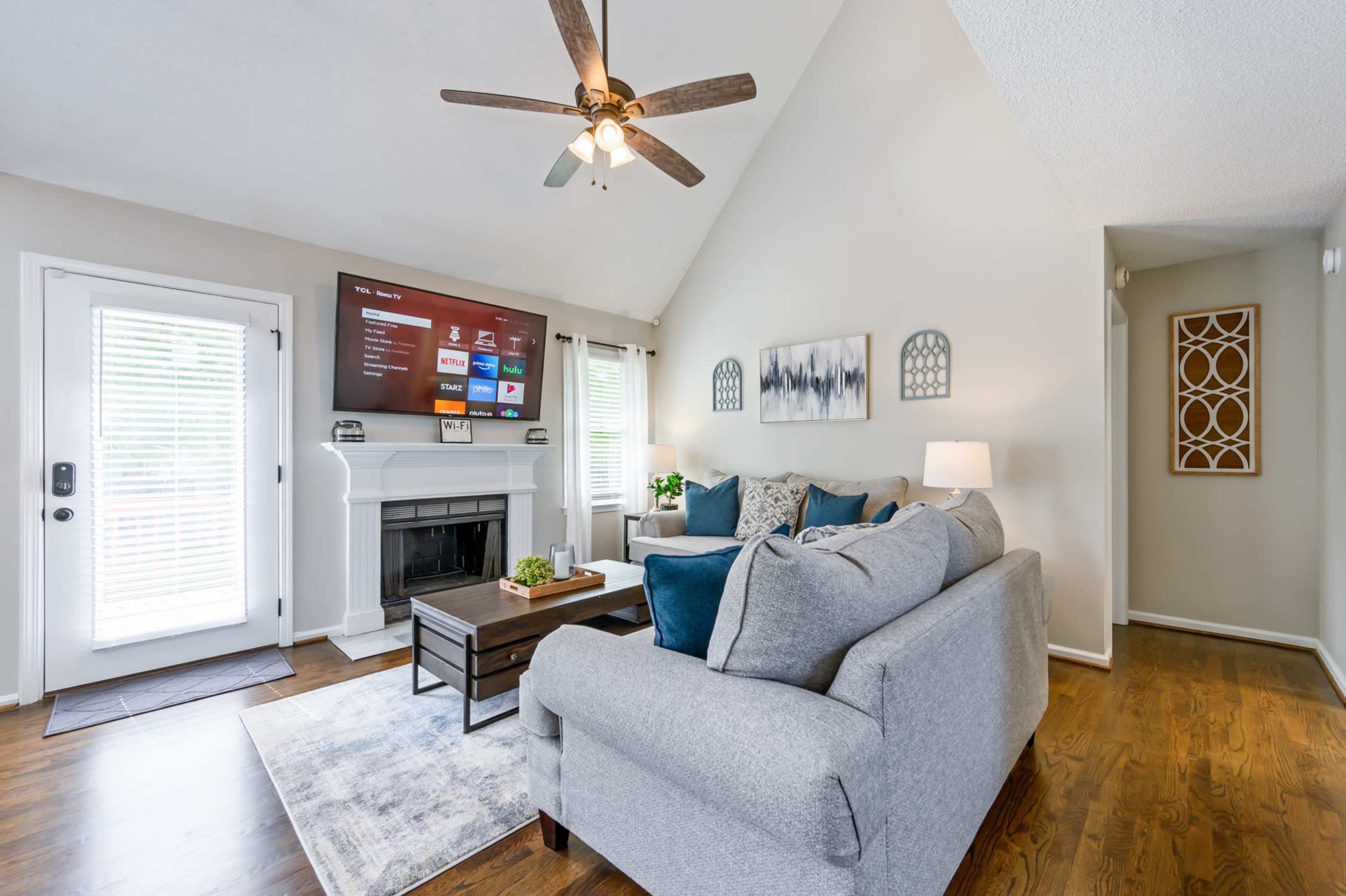 A living room features a gray sectional sofa, a wooden coffee table, and a mounted television above a fireplace, all illuminated by natural light.