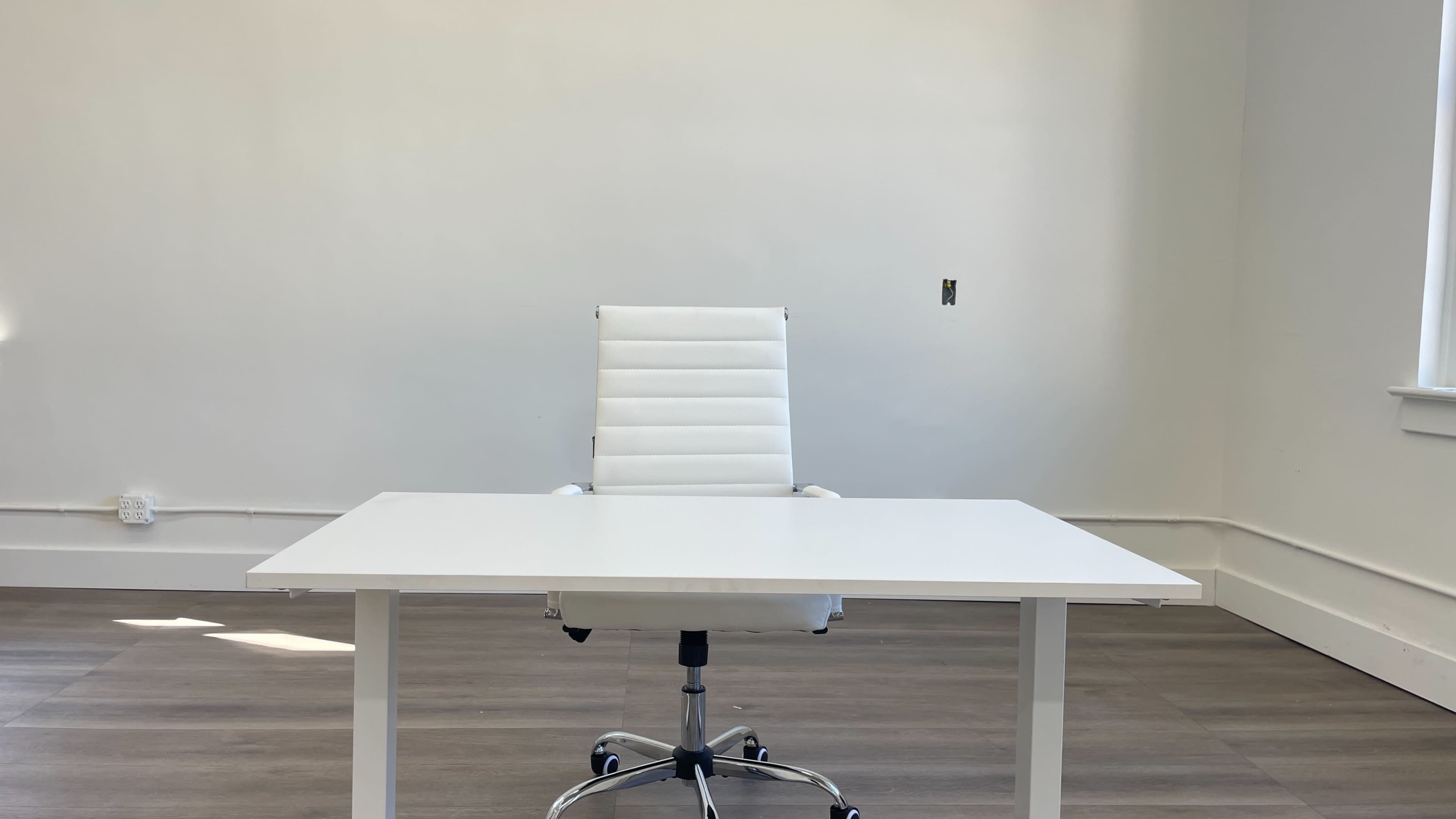 A white desk and a white chair are positioned in a minimally furnished room with light wood flooring and plain walls.