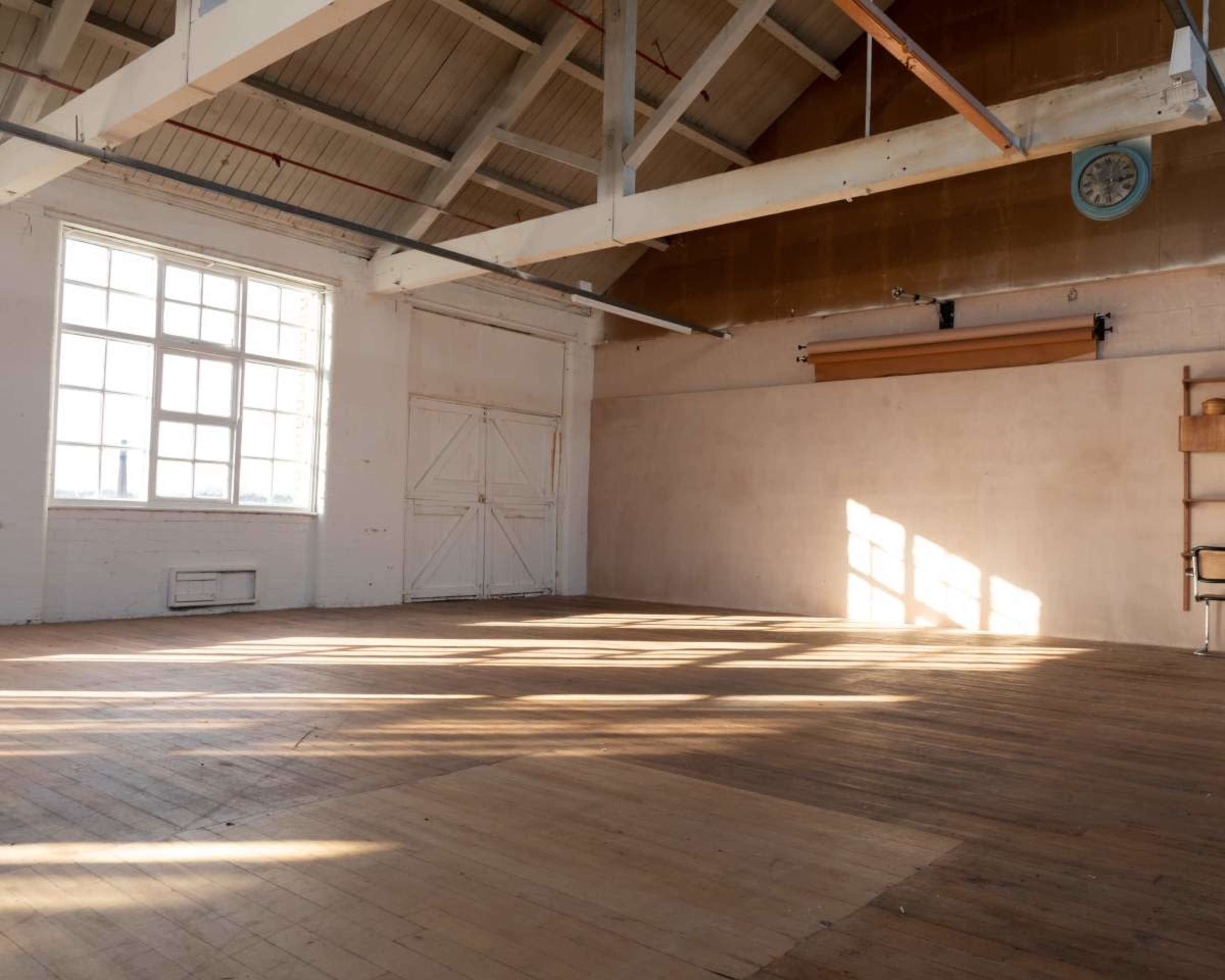 The image shows an empty room with wooden flooring, large windows allowing natural light to stream in, and a minimalist aesthetic.