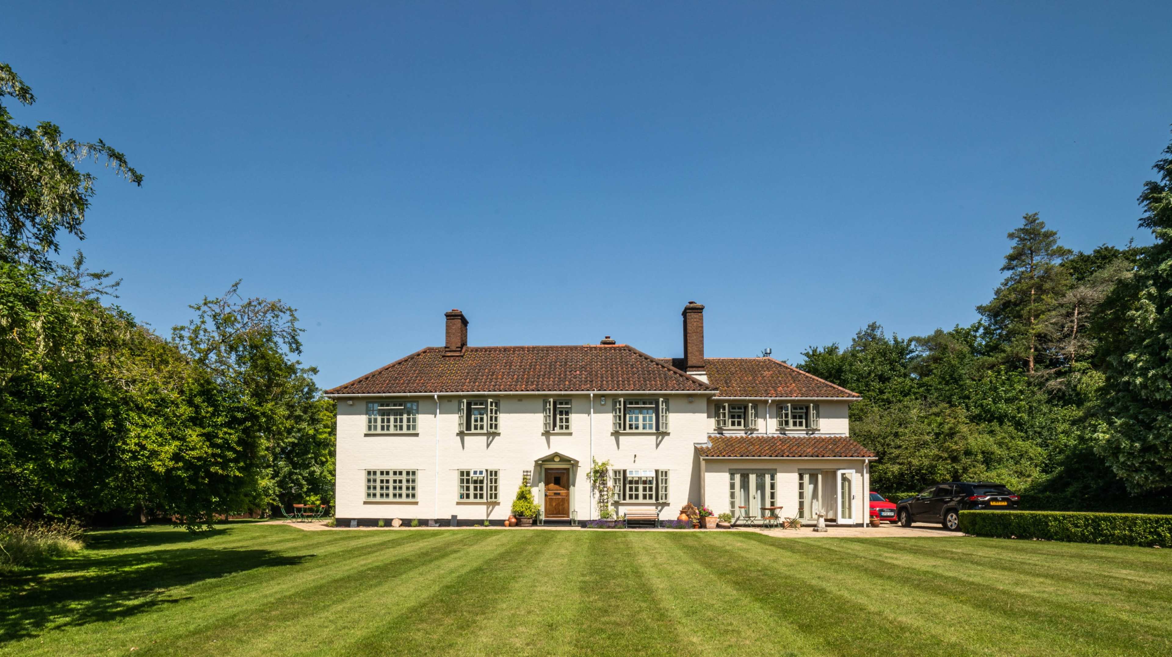 A two-story white house with a tiled roof sits on a well-maintained lawn surrounded by trees.