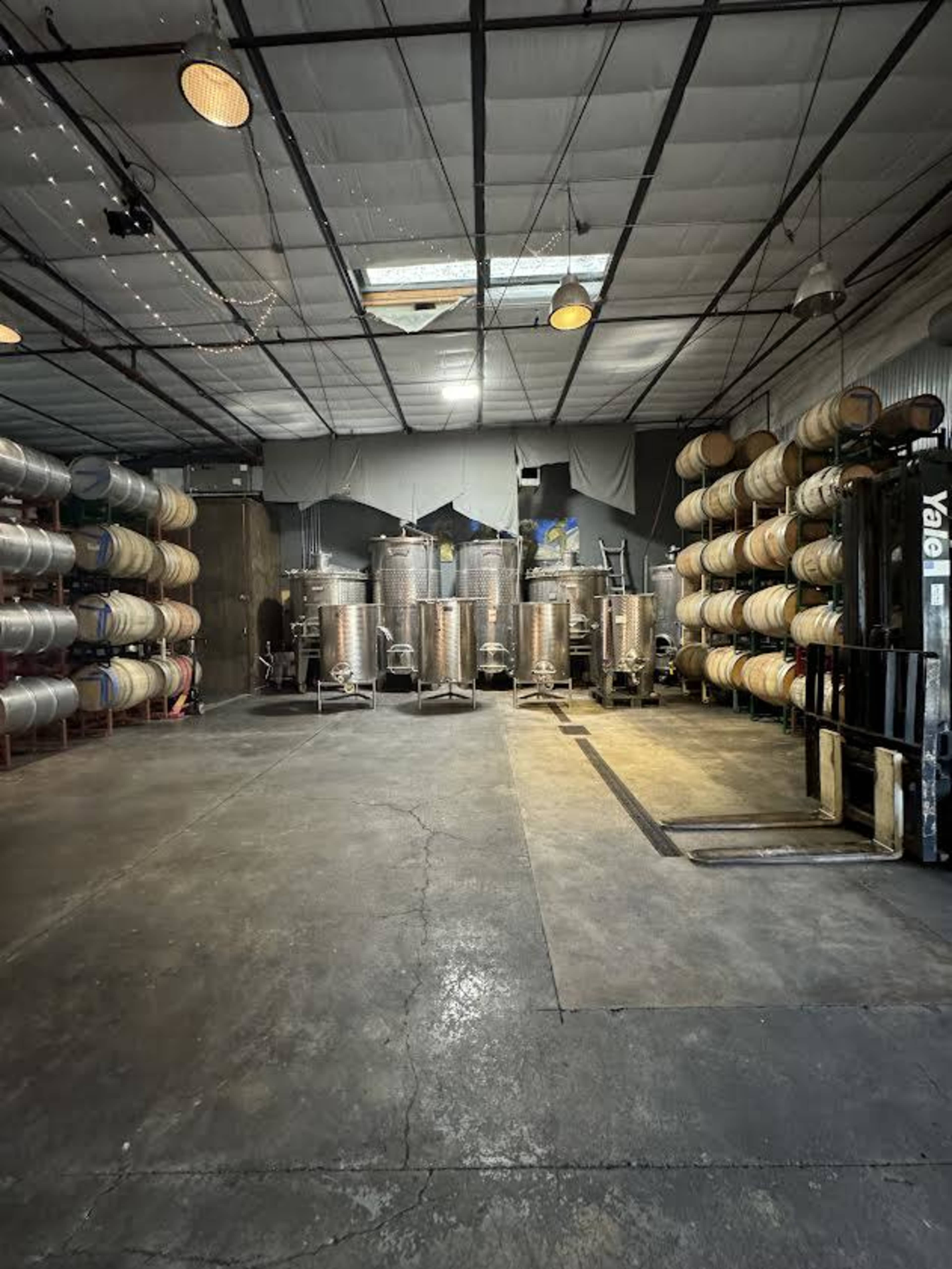 An industrial interior storage space with stacked barrels on either side and metal fermentation tanks arranged in the center.