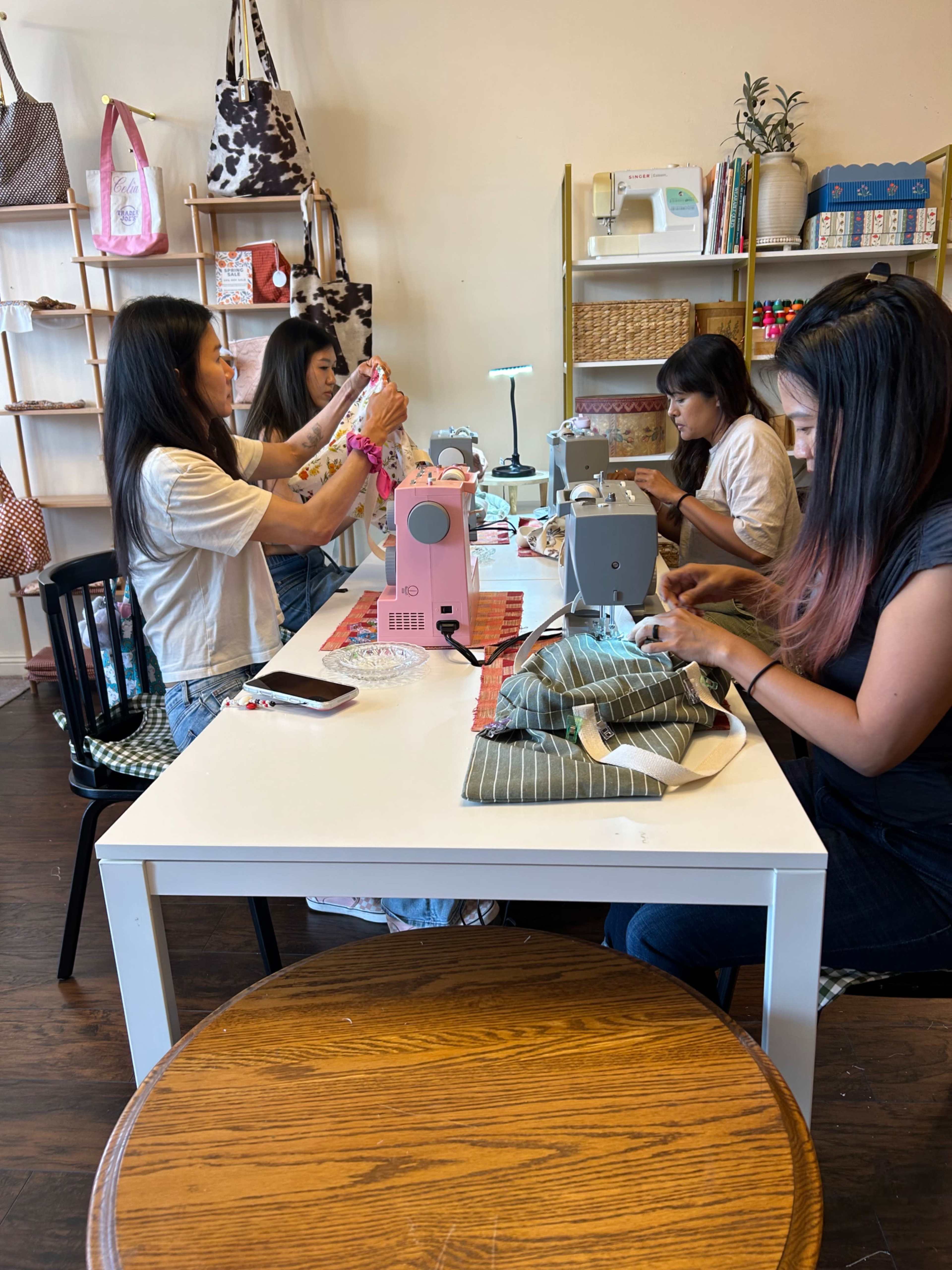Four individuals are seated at a table engaged in a sewing workshop, working on various fabric projects with sewing machines.