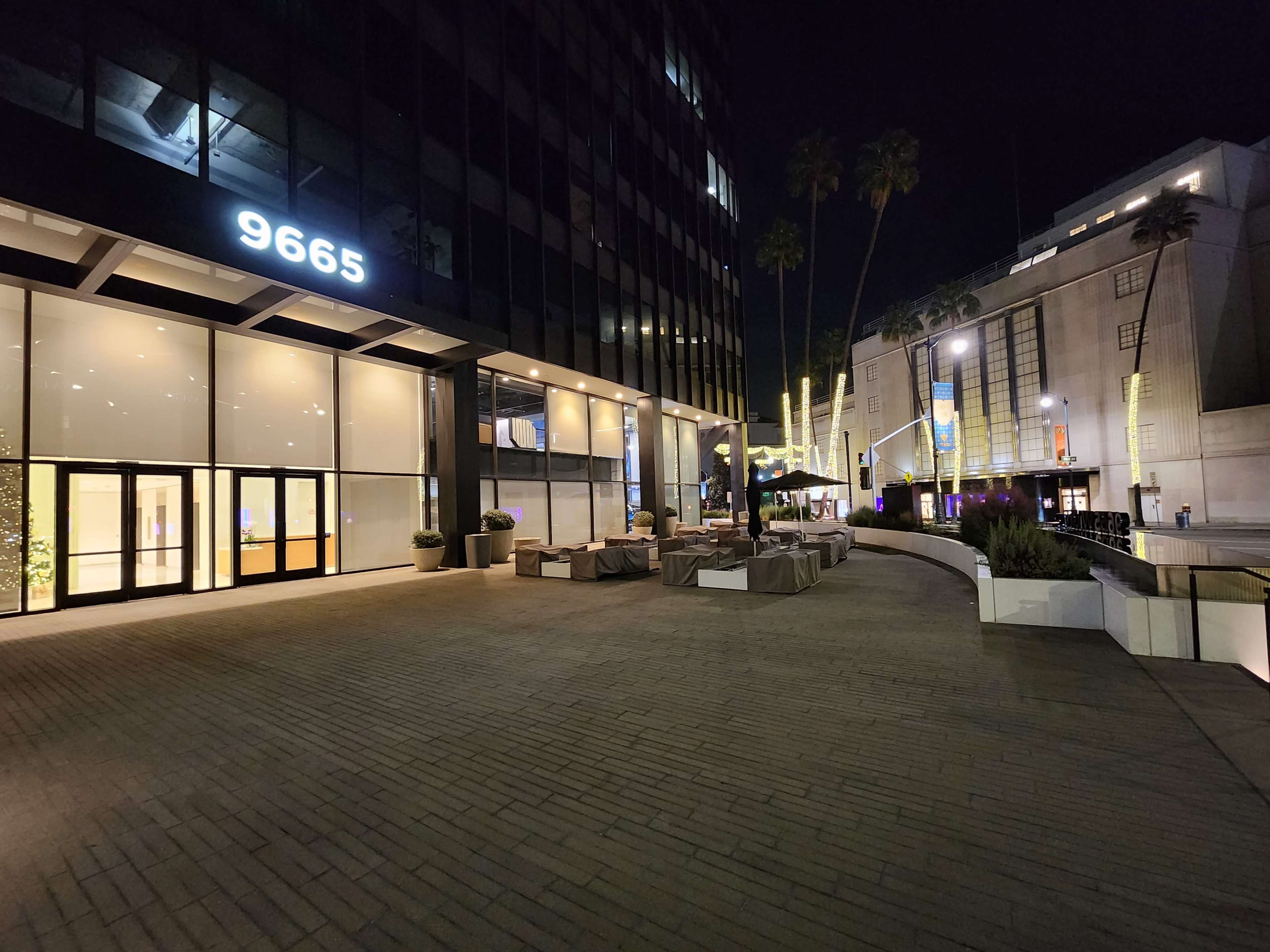 A modern building at night with the number "9665" displayed prominently, surrounded by outdoor seating and palm trees.