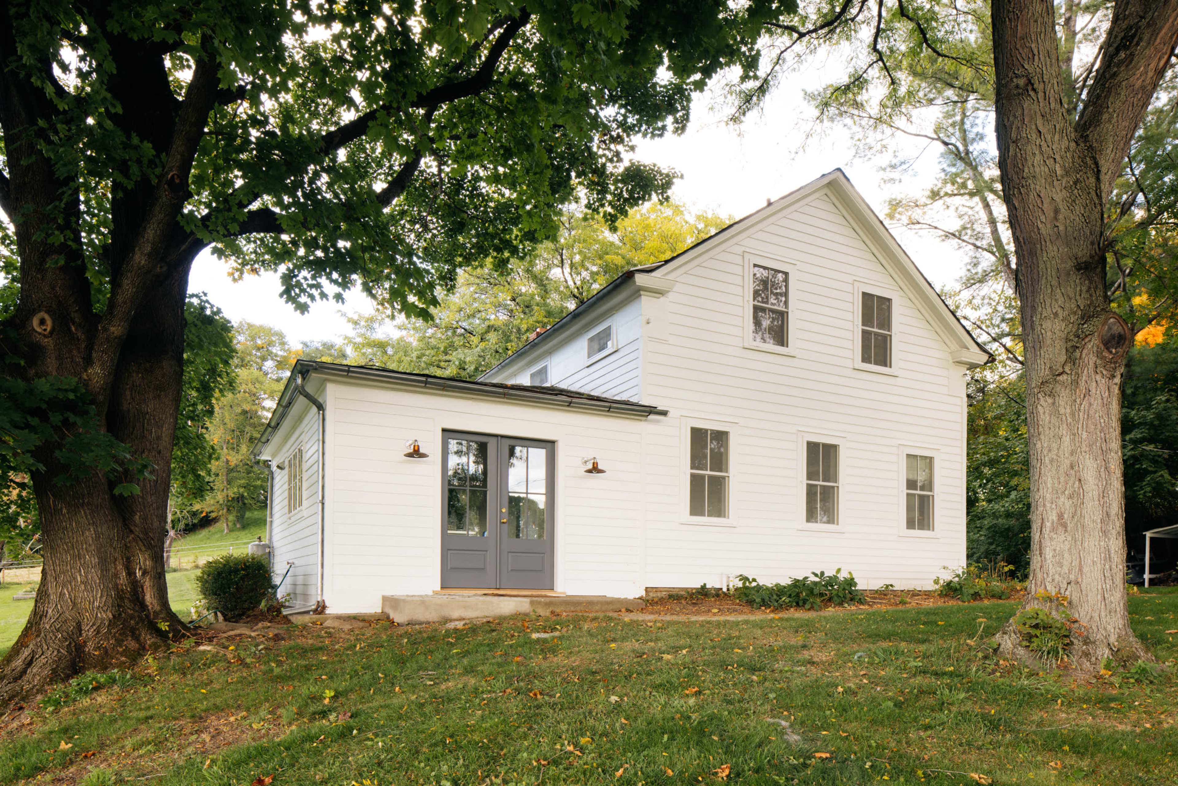 A white two-story house with gray doors and windows, surrounded by green grass and trees.