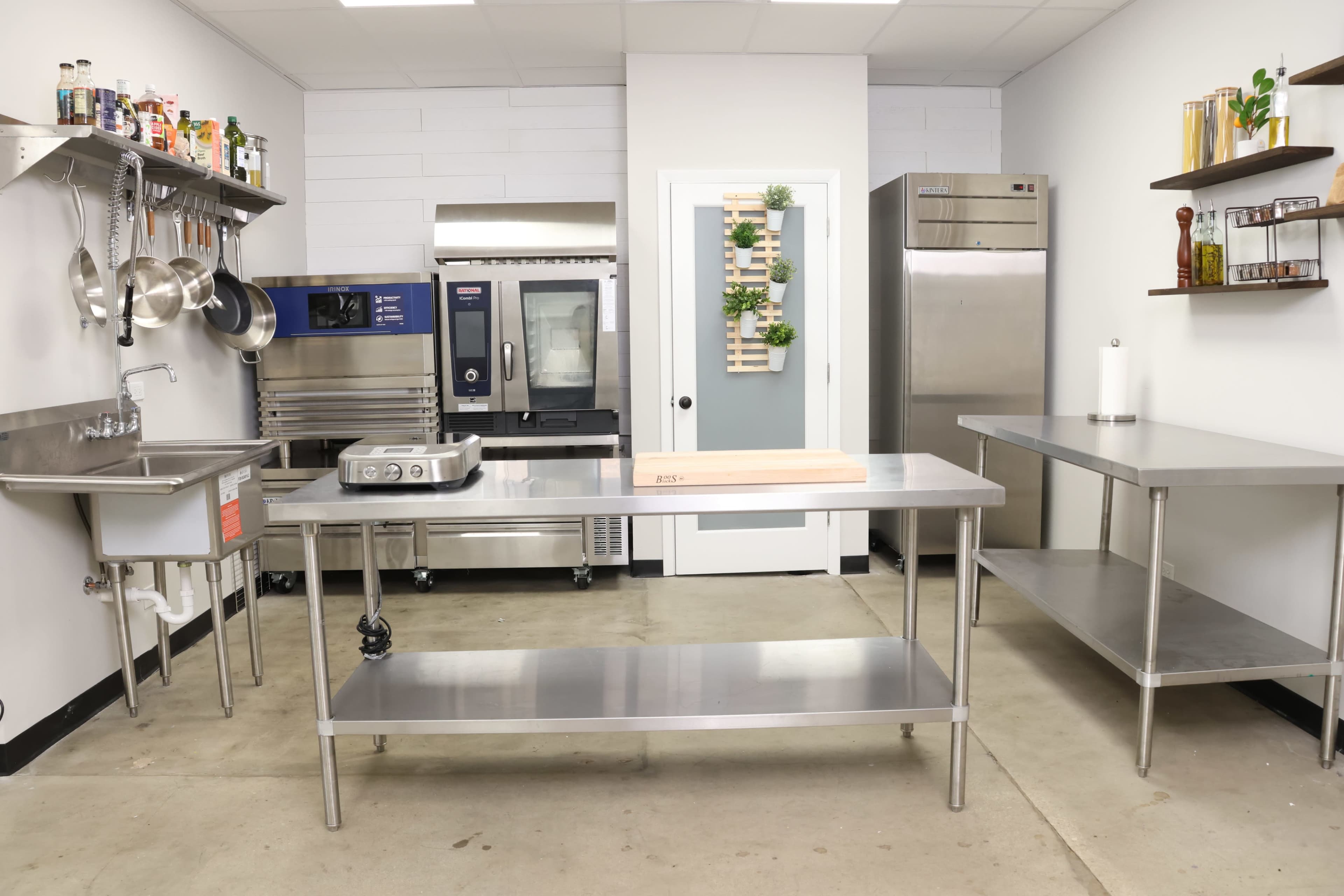 A modern commercial kitchen with stainless steel countertops, a sink, and various kitchen appliances including a refrigerator and an oven.