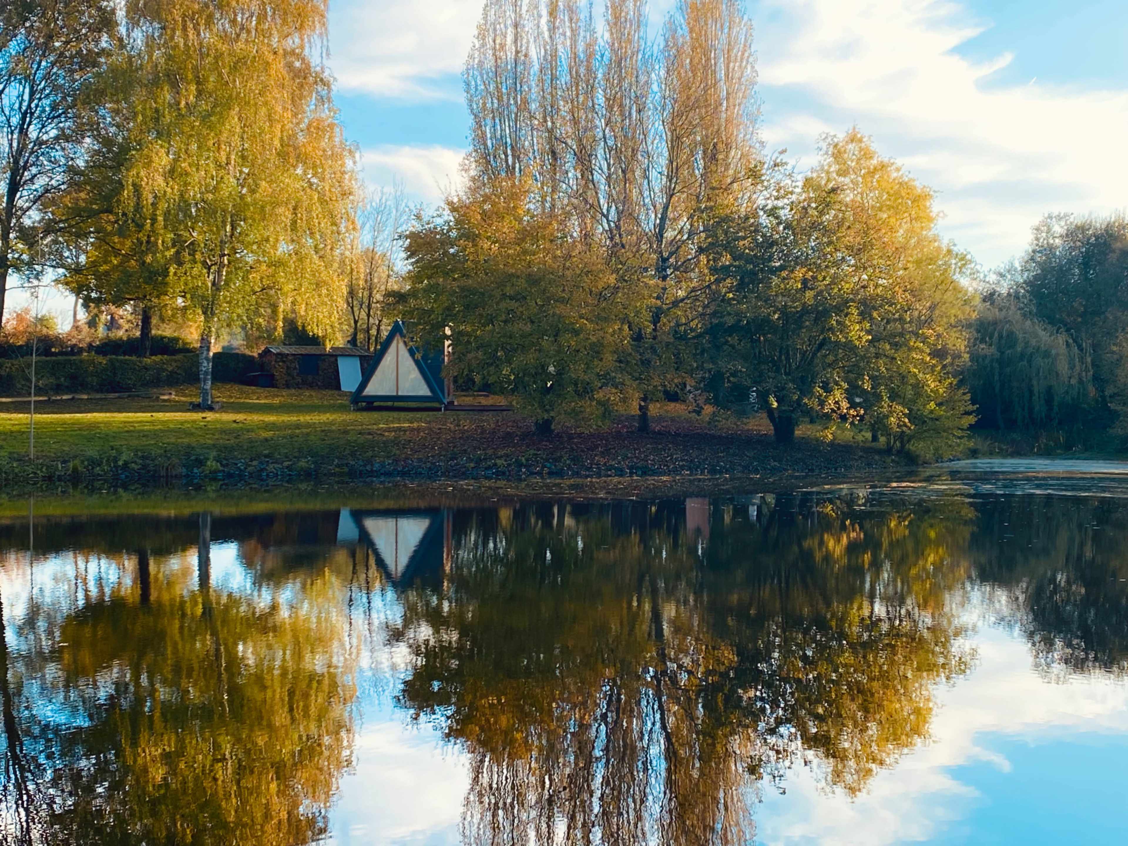 A triangular structure is positioned by a pond surrounded by trees displaying autumn foliage.