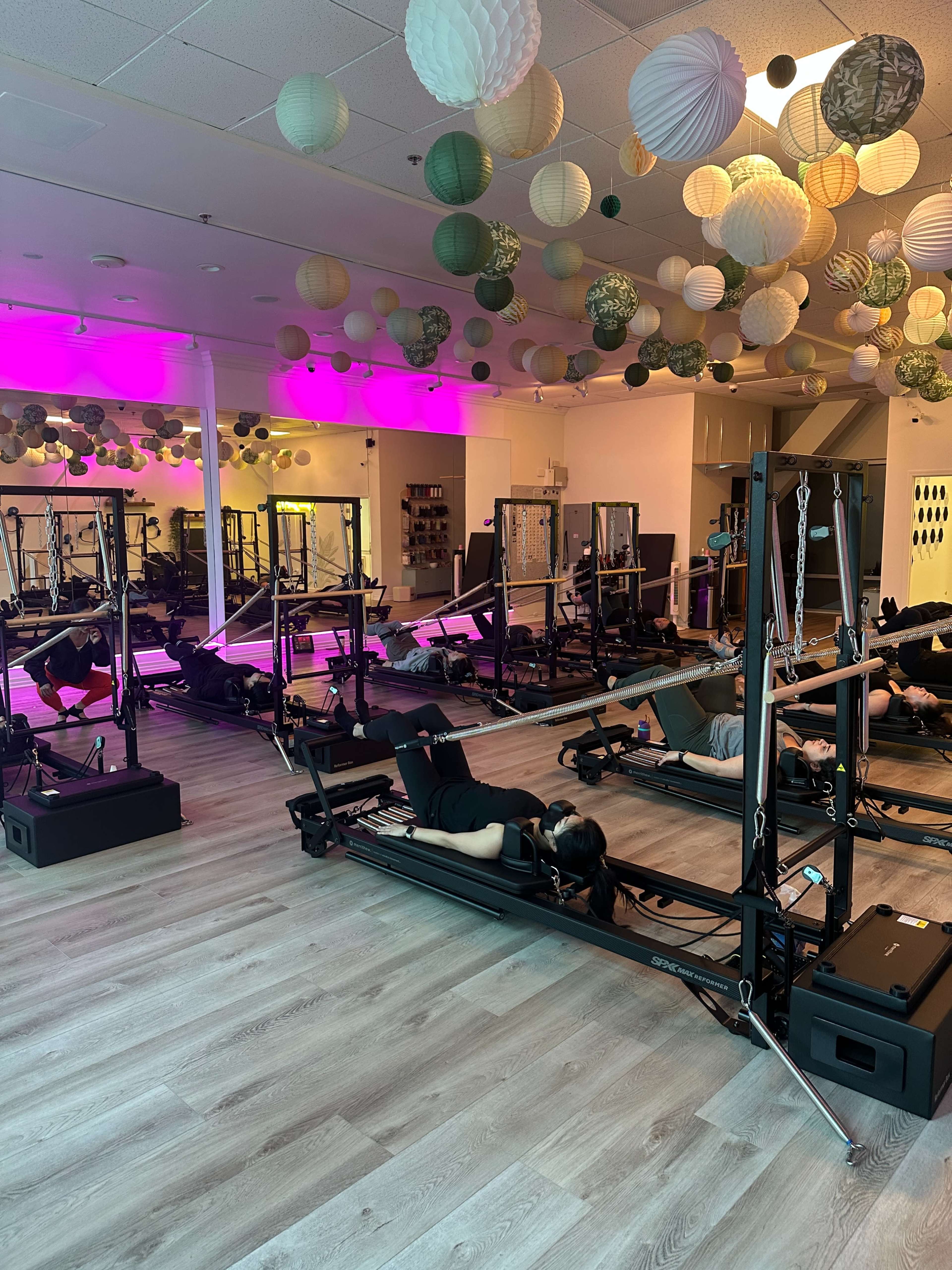 The image shows a fitness studio with multiple reformer machines arranged neatly, featuring colorful paper lanterns hanging from the ceiling and soft lighting.