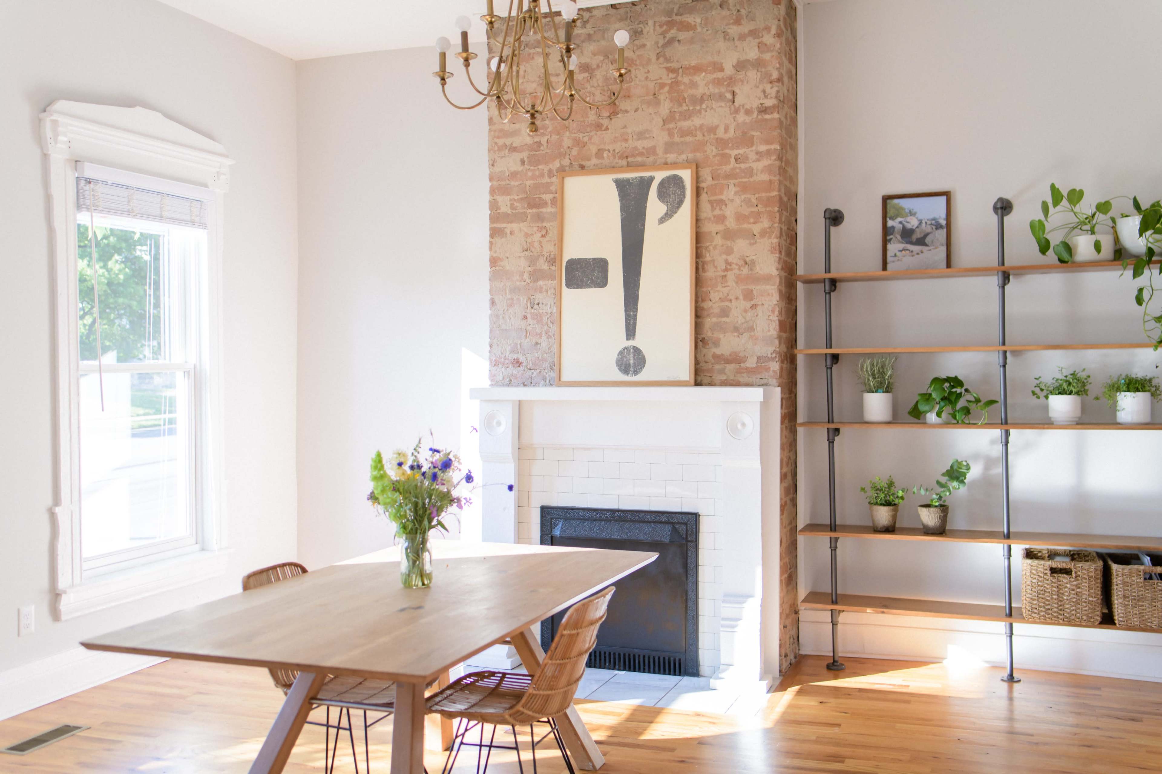 A cozy dining area features a wooden table surrounded by chairs, a fireplace with a white mantel, exposed brick wall, and a minimalist art piece above the fireplace, complemented by a shelf of plants.
