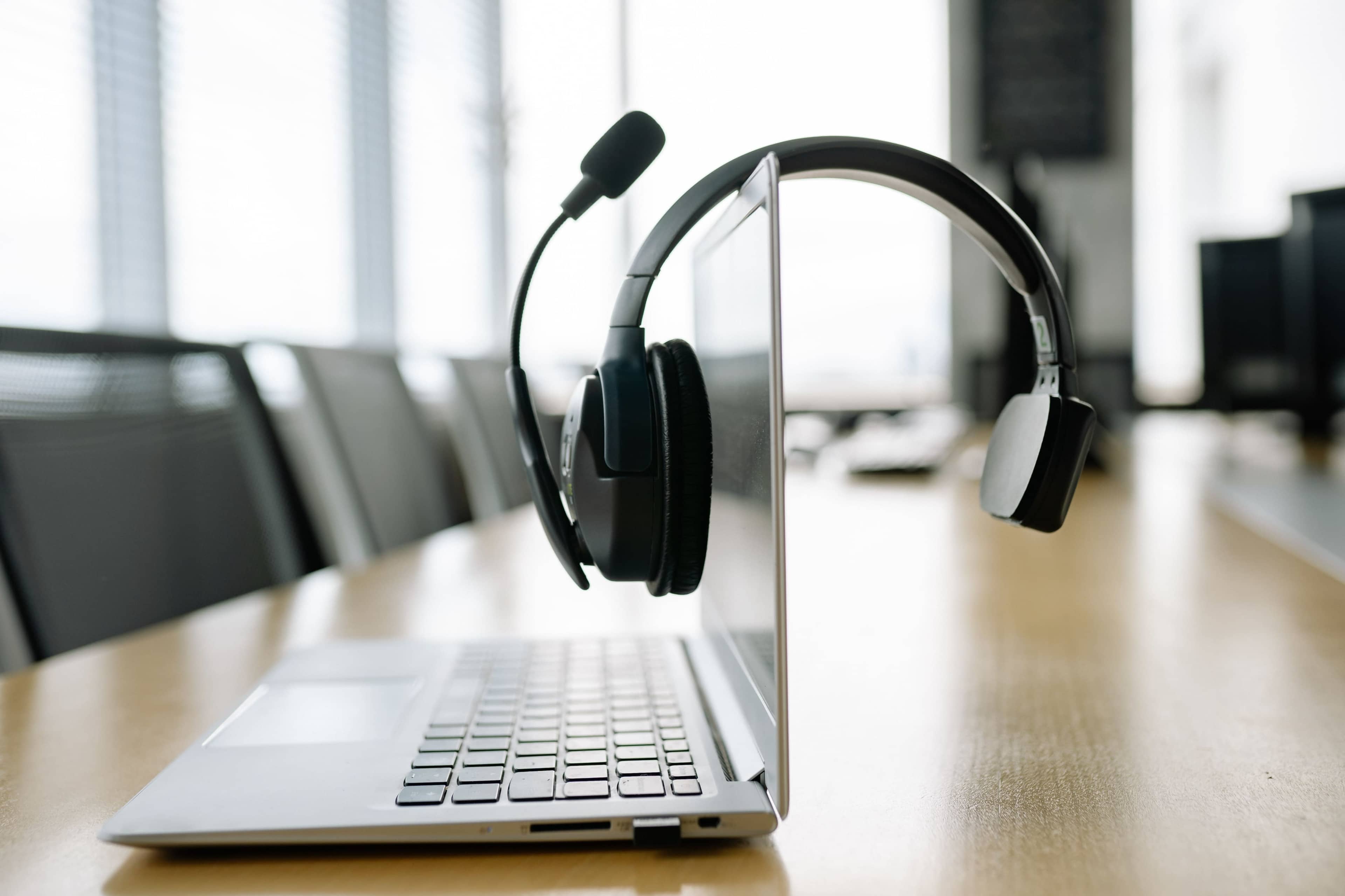 A black headset rests on the edge of a silver laptop on a wooden table in a modern office setting.