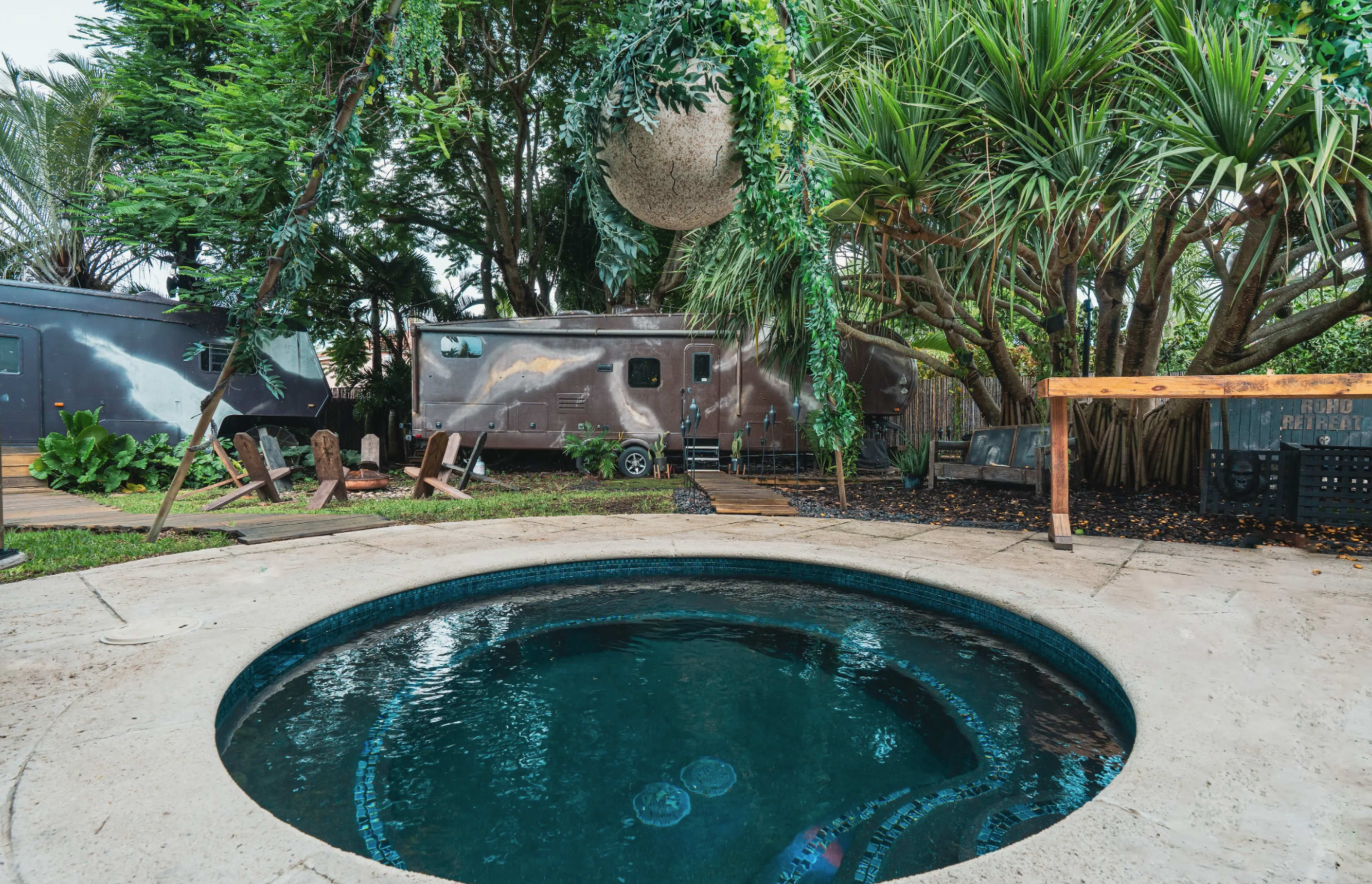 The image shows a circular hot tub surrounded by a lush garden with palm trees and trailers in the background.