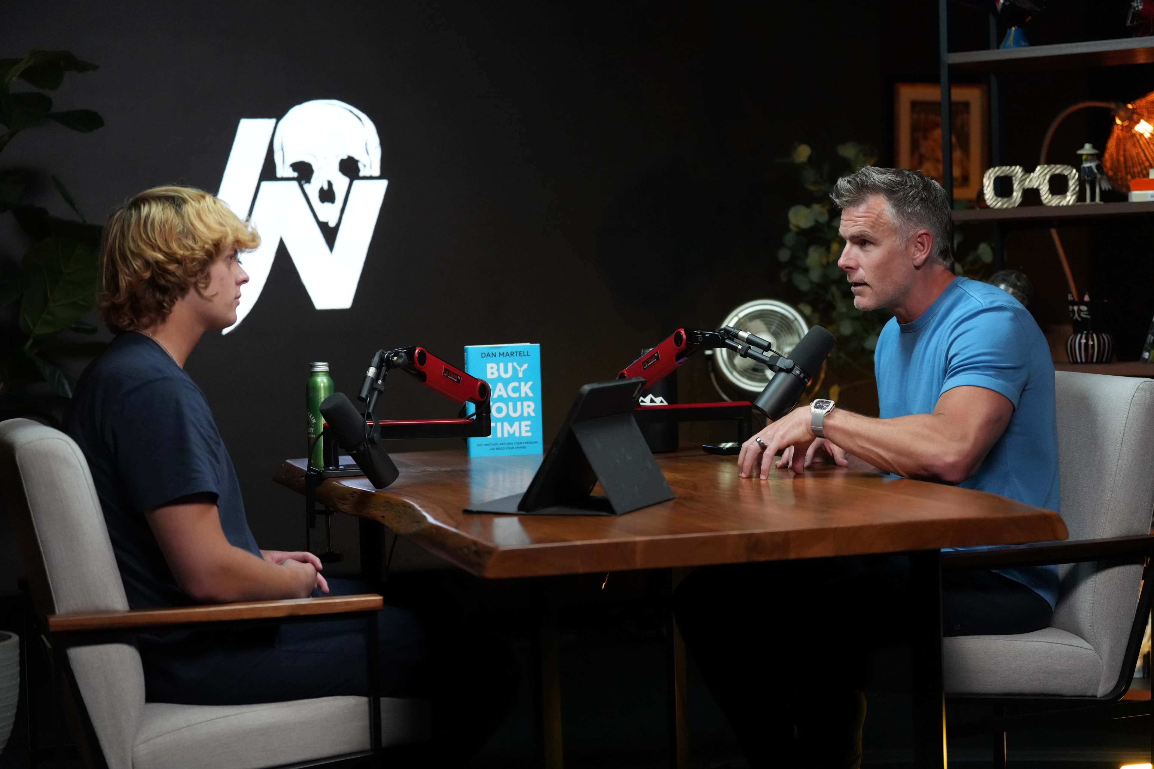 A man in a blue shirt is discussing a book titled "Buy Back Our Time" with a young man seated at a wooden table in a studio setting.