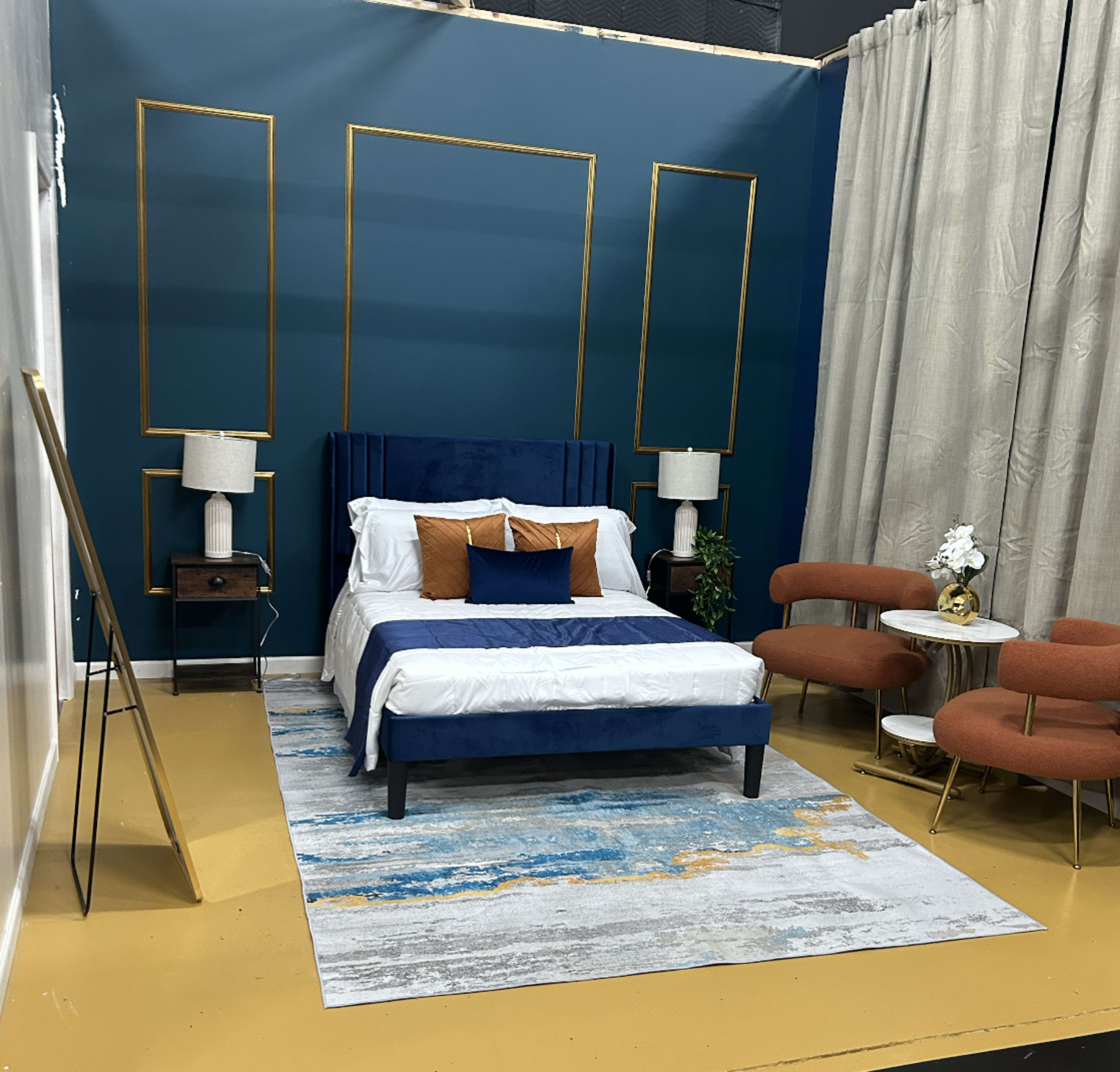 The image shows a styled bedroom featuring a blue upholstered bed, orange accent pillows, two orange lounge chairs, and a round coffee table on a patterned rug, all set against a teal wall with gold frames.