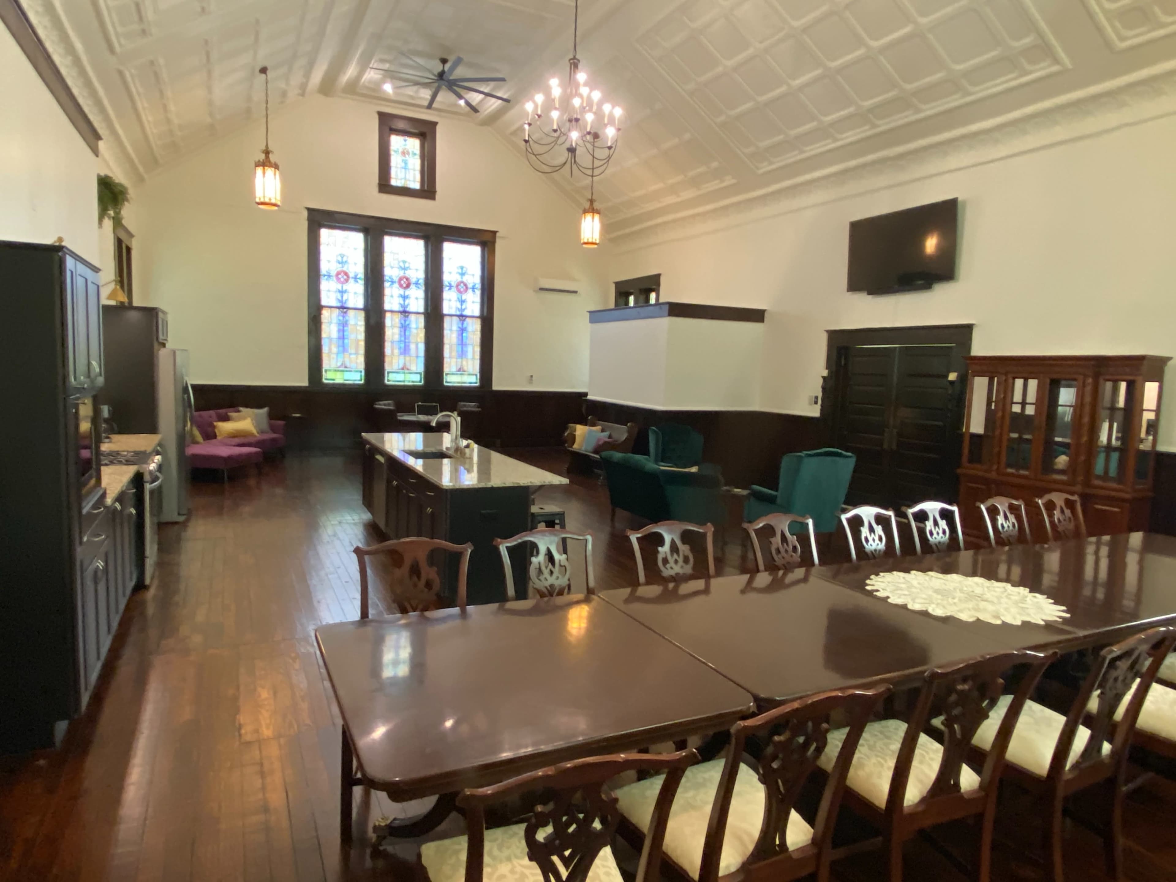 The image shows a spacious room with high ceilings, stained glass windows, and a long dining table surrounded by chairs, alongside a kitchen area and seating arrangements.