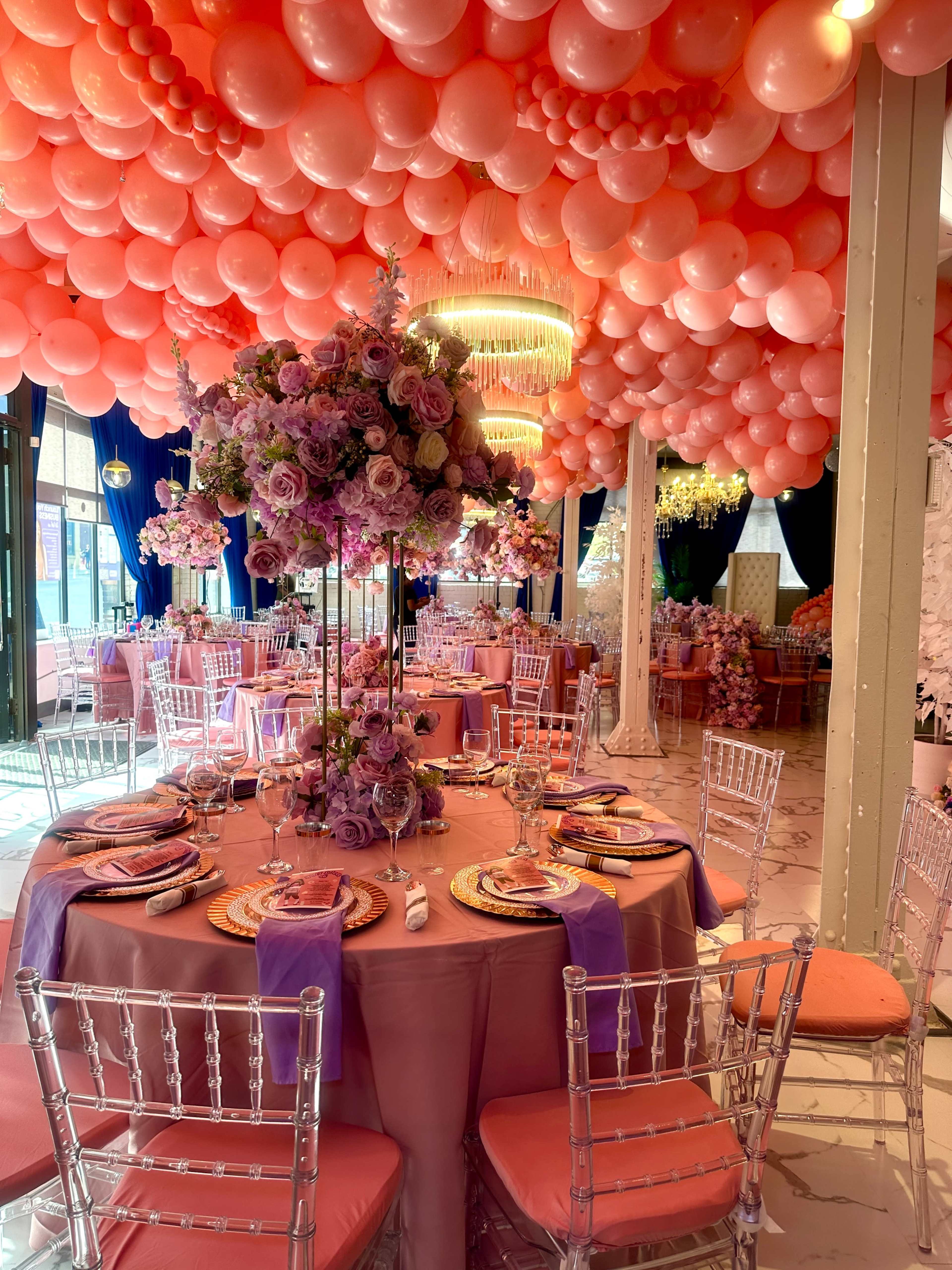 The image features a decorated event space with tables set for dining, surrounded by a ceiling adorned with pink balloons and floral centerpieces.