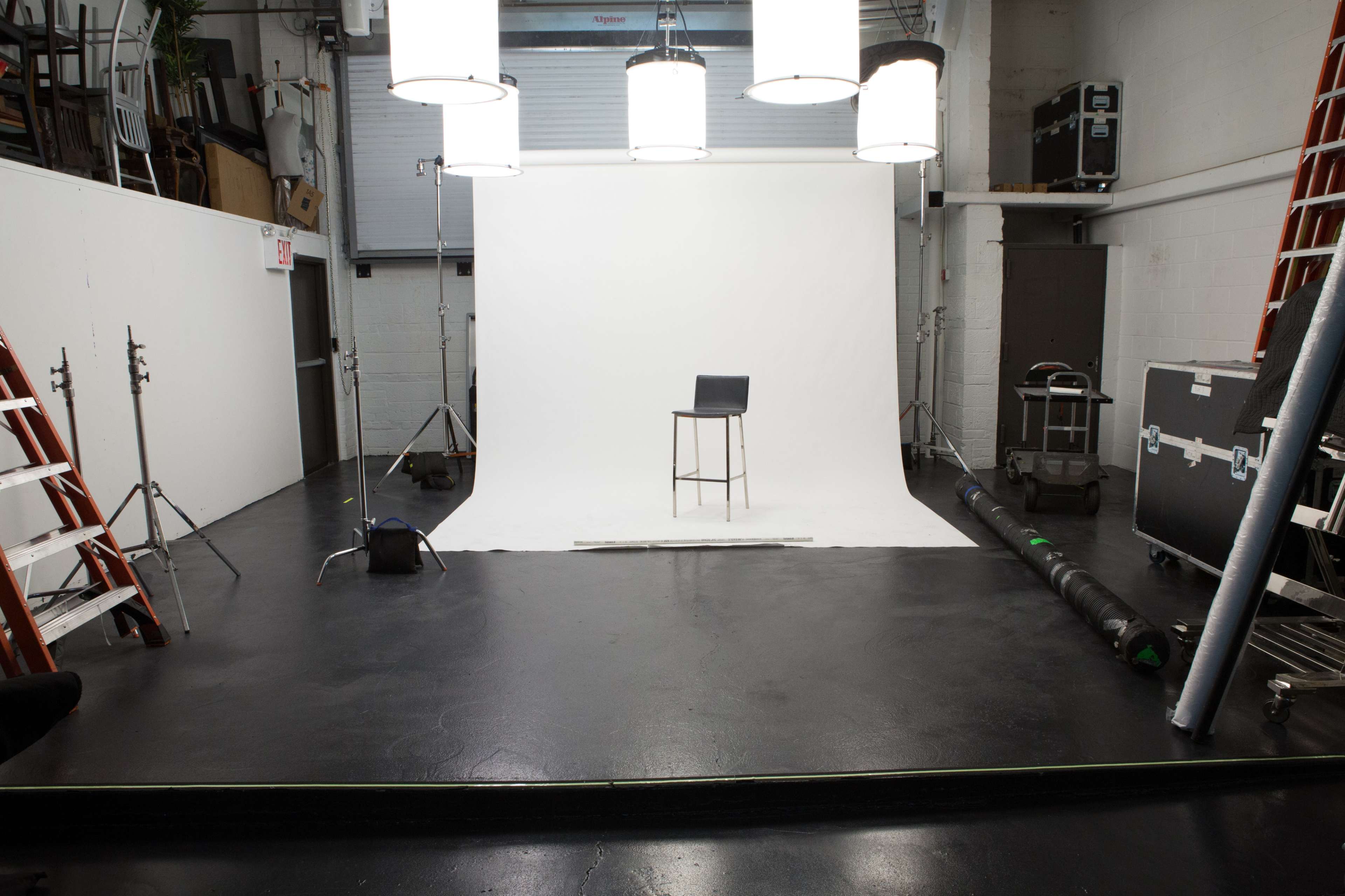 A photography studio with a white backdrop, a tall stool in the center, and several light fixtures hanging above, along with equipment and ladders positioned around the space.