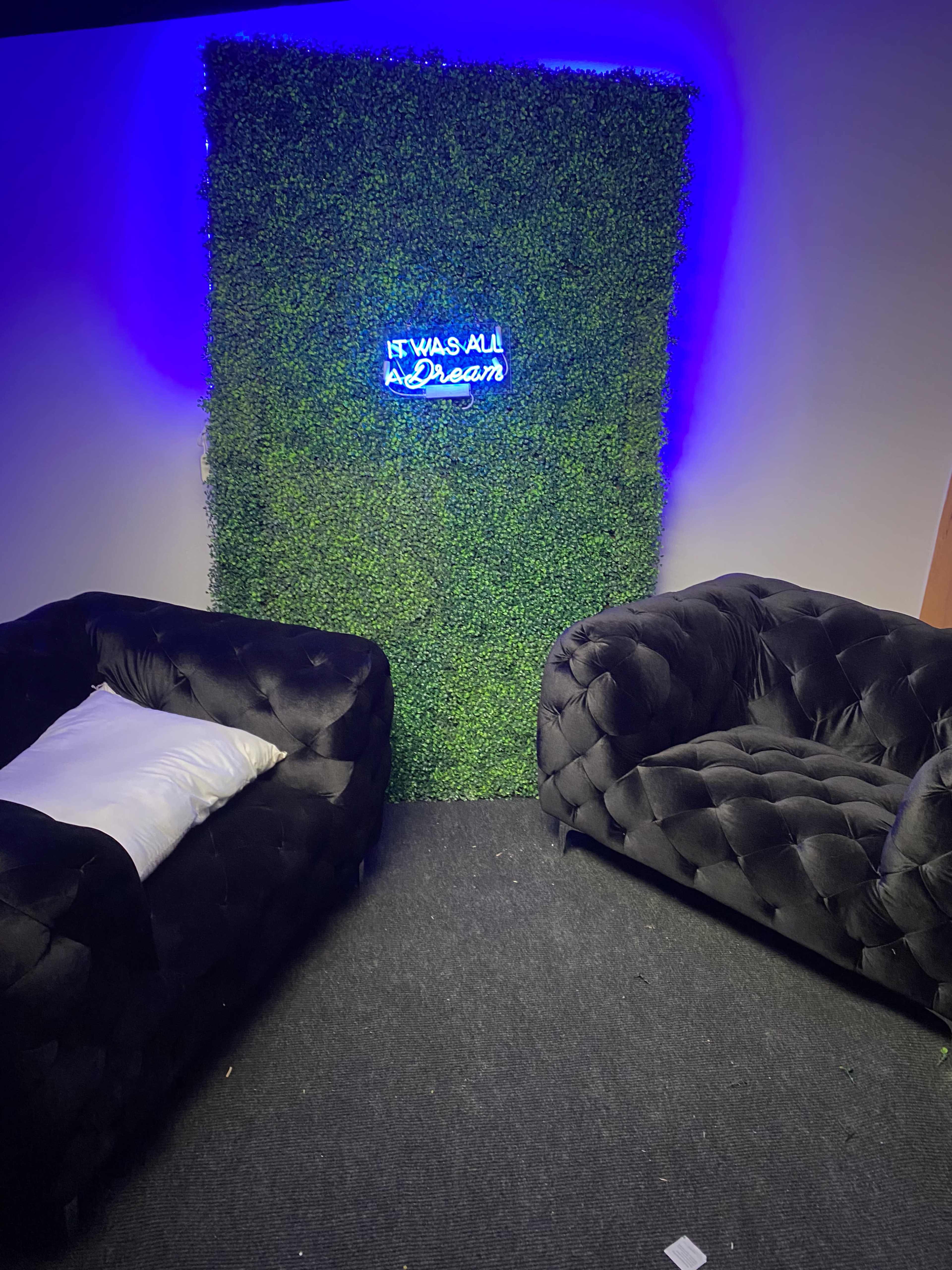 The image shows a small indoor space featuring two black tufted sofas facing a vertical green wall with a neon sign that reads, "IF IT WAS ALL A DREAM."